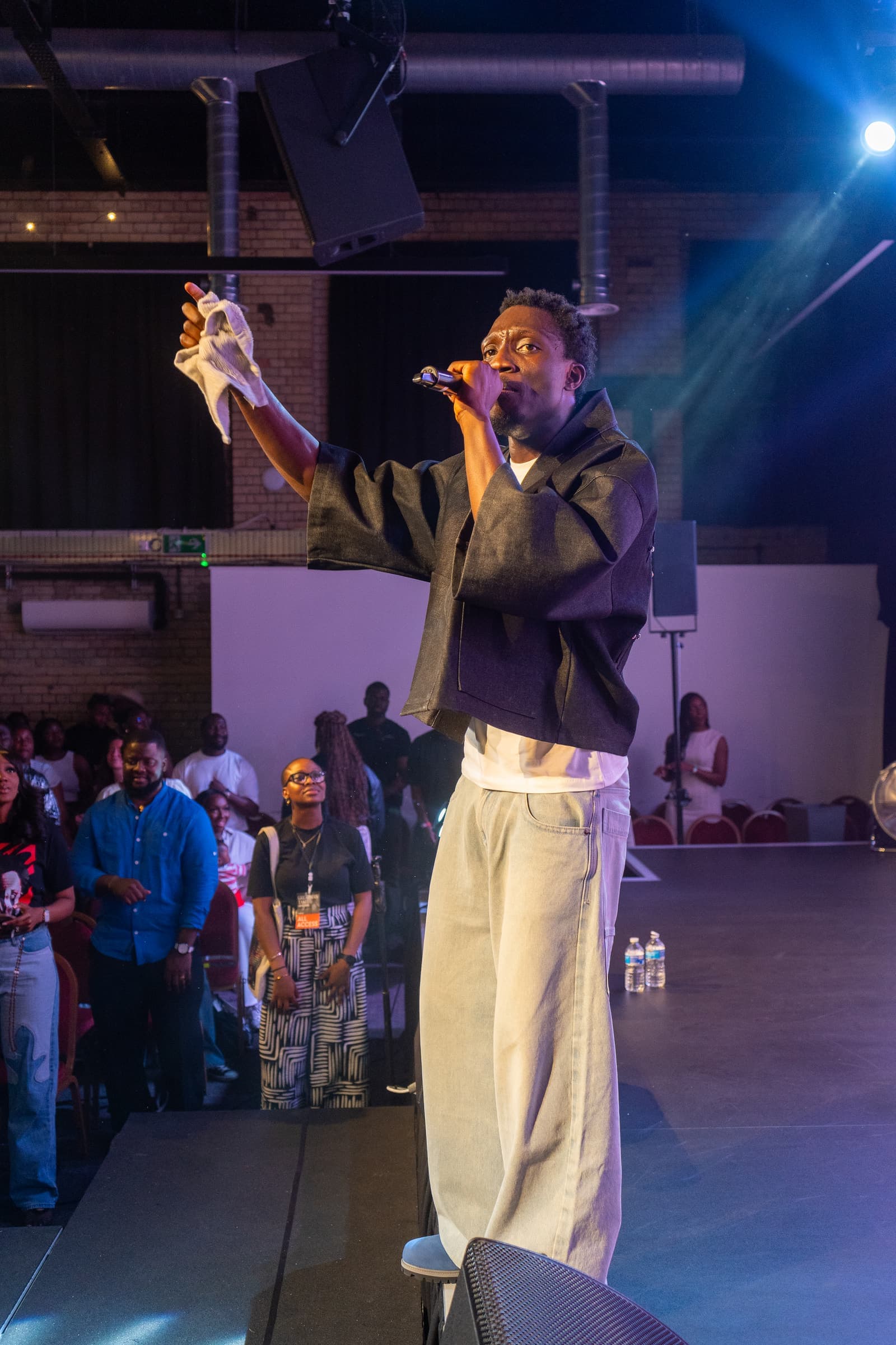 A performer mid-verse under the stage lights, one hand raised with a white towel and the other holding the mic, the front of the room on its feet behind him.