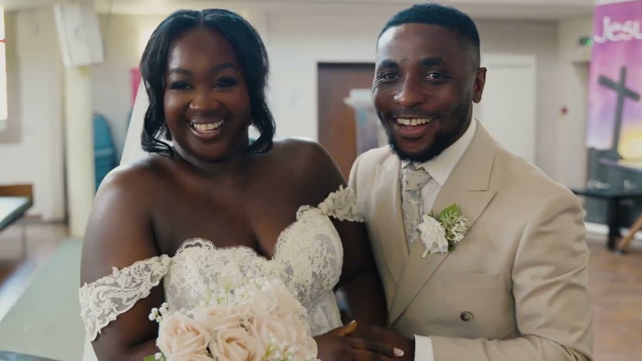 The couple side by side to camera, both laughing — she in an off-shoulder lace gown holding blush roses, he in a cream suit with a white boutonniere.
