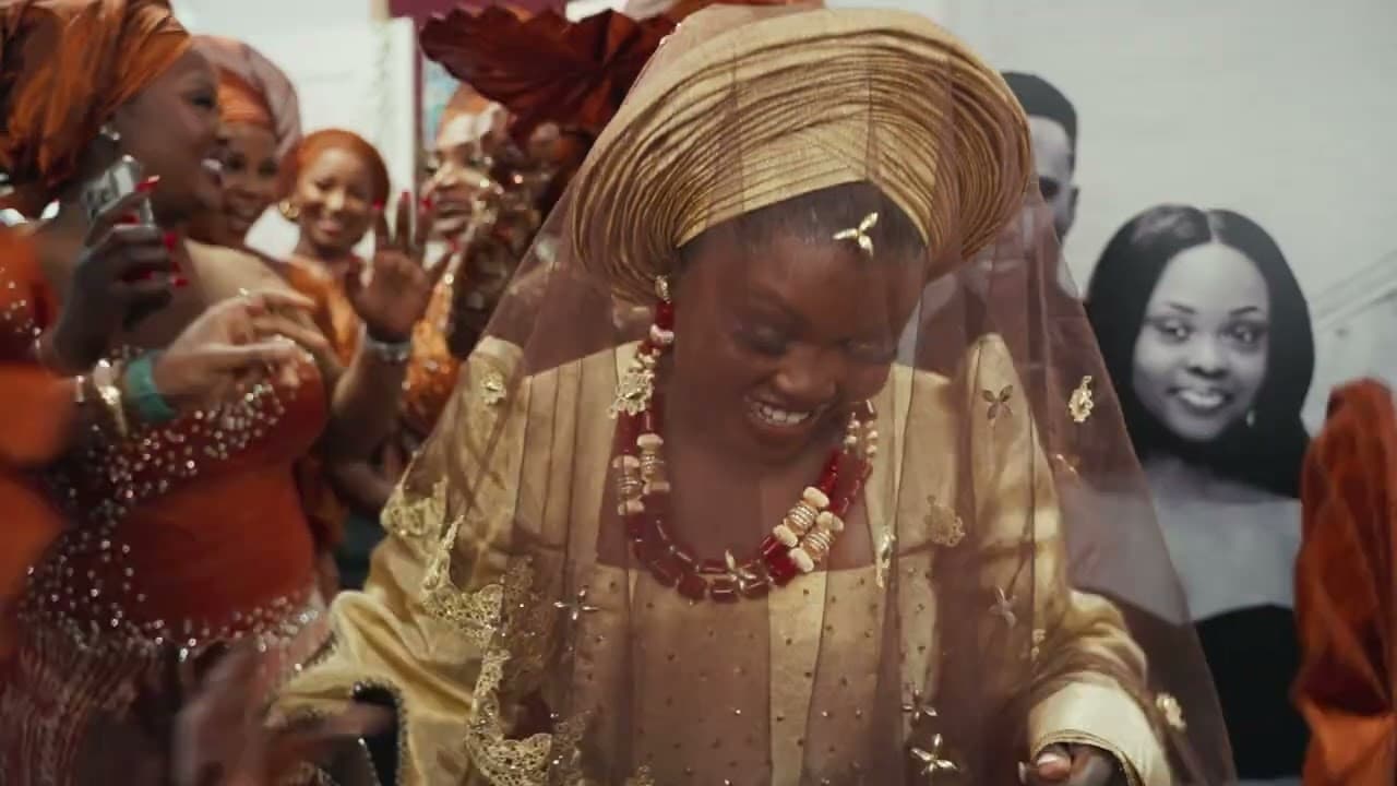 The bride in a gold aso-oke gele and beaded coral necklace, laughing under a sheer veil as her party dance around her in burnt orange.