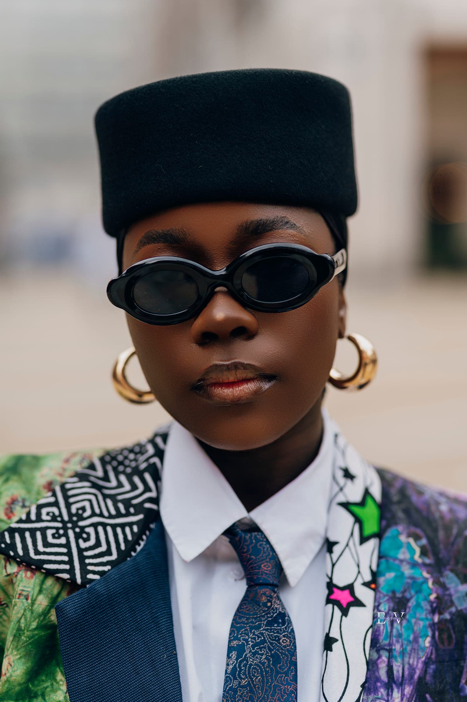 A straight-on portrait of a woman in a black pillbox hat and black oval sunglasses, gold hoop earrings, a white collar and a blue paisley tie under a patchwork jacket.