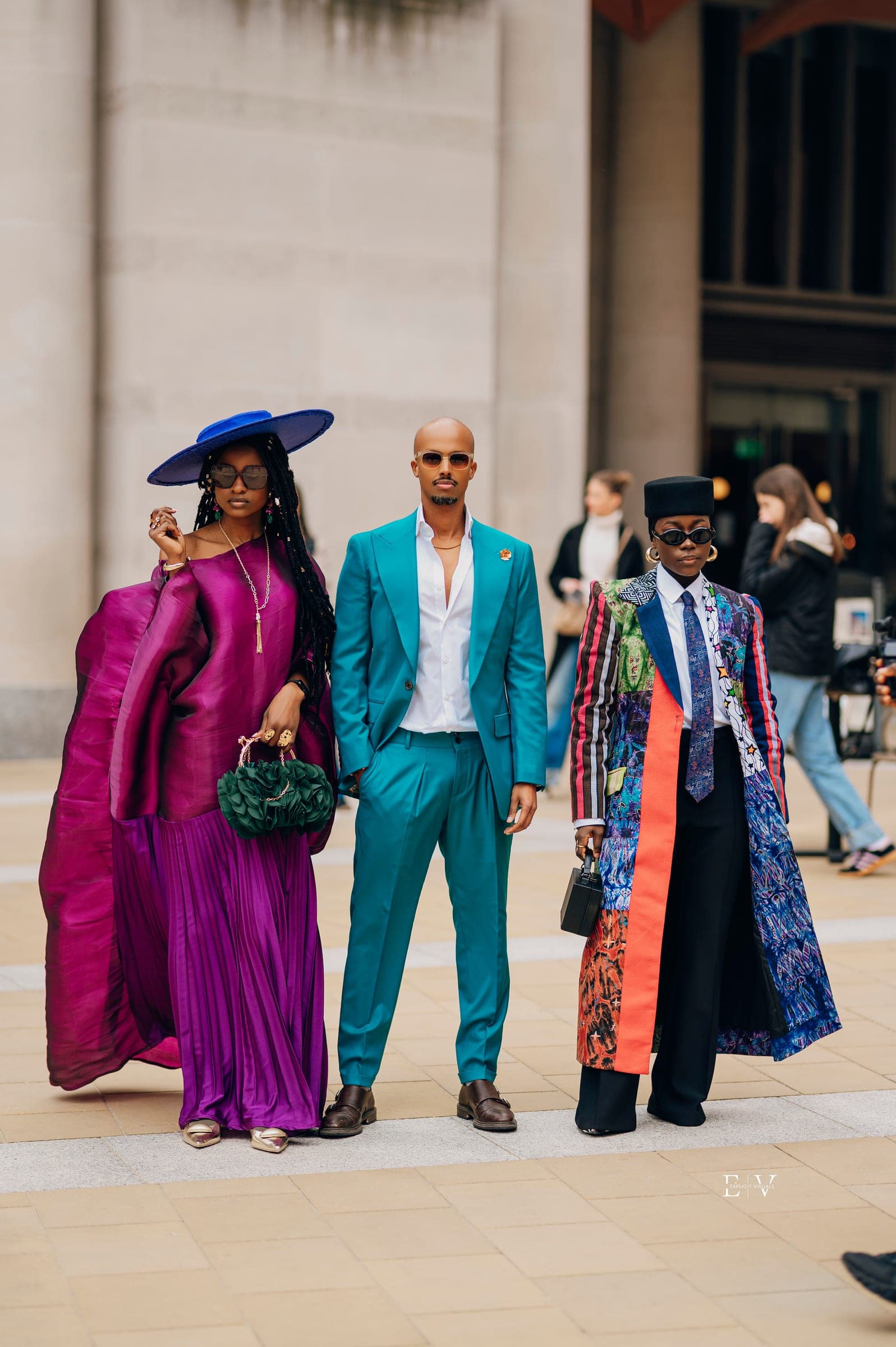 Three guests together — a woman in a magenta pleated gown and a wide blue hat, a man in a teal satin suit, and a guest in a printed long coat and black cap.