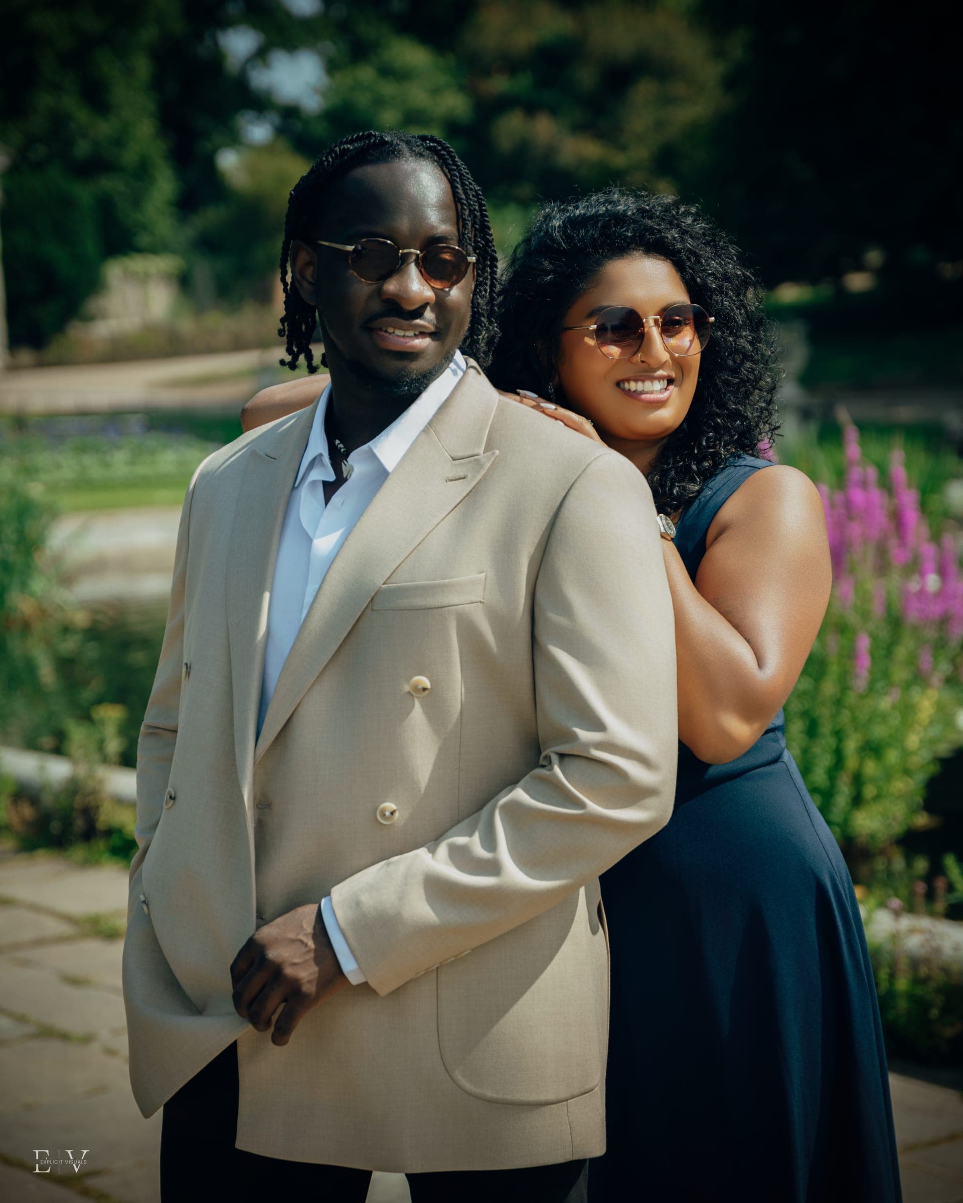Pahi and Eugene in a sunlit park garden, arms around each other in a cream double-breasted blazer and a navy dress, both smiling behind sunglasses.