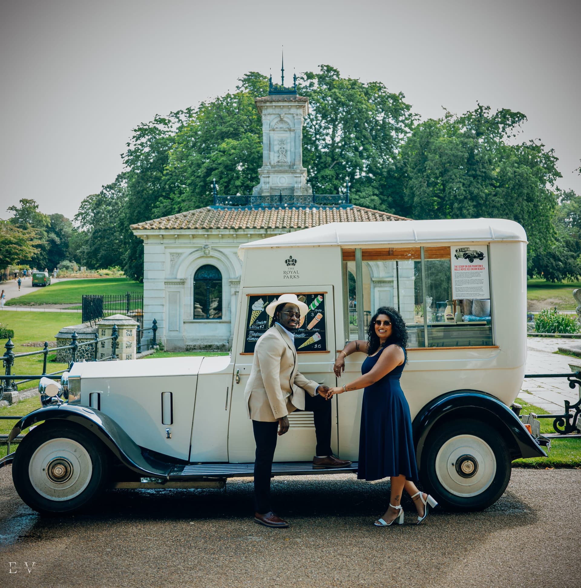 Standing either side of the van's open door with hands linked across it, a domed stone monument and a garden pavilion behind them.
