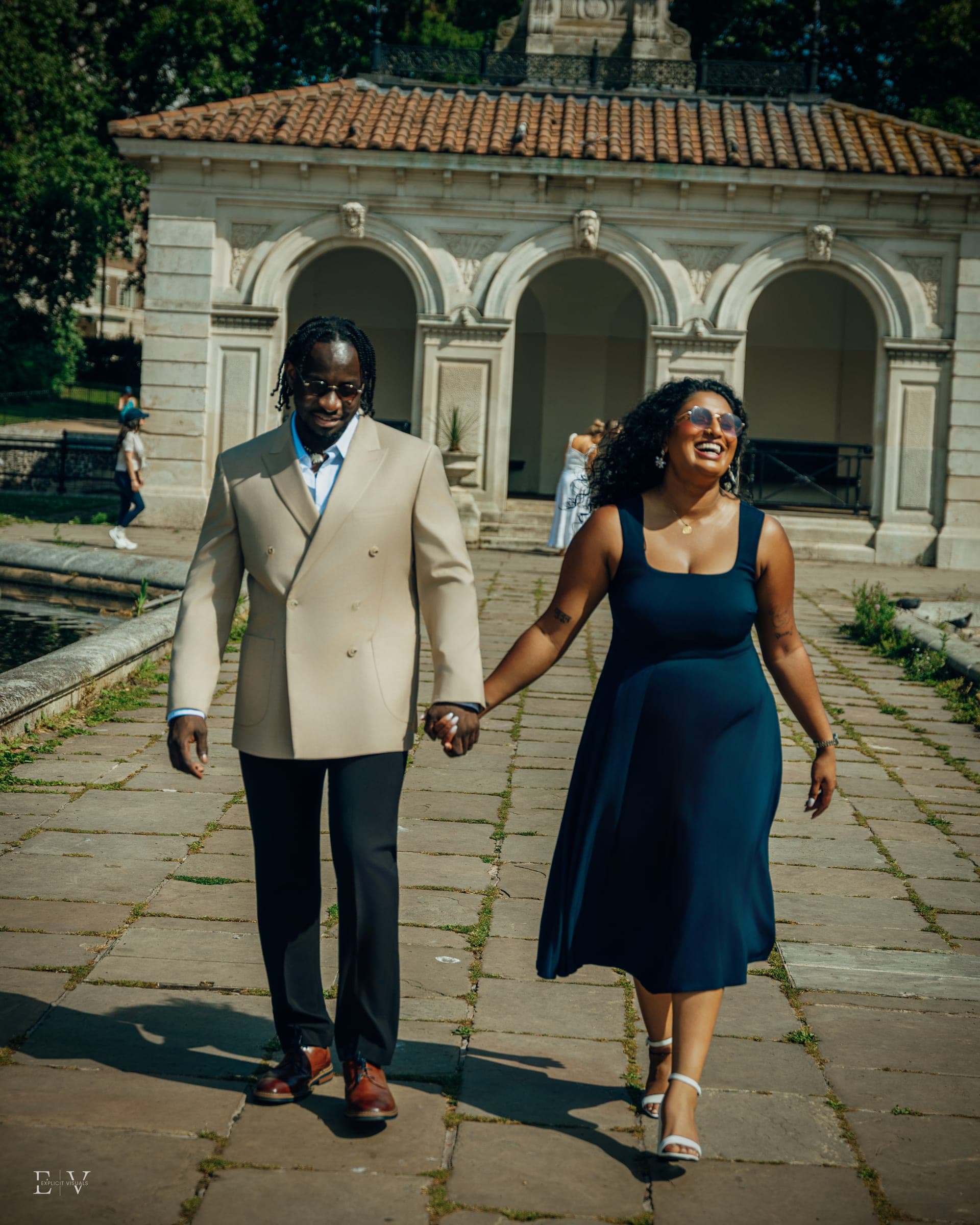 Walking hand in hand along a stone path toward the camera, an arcaded garden pavilion behind them.