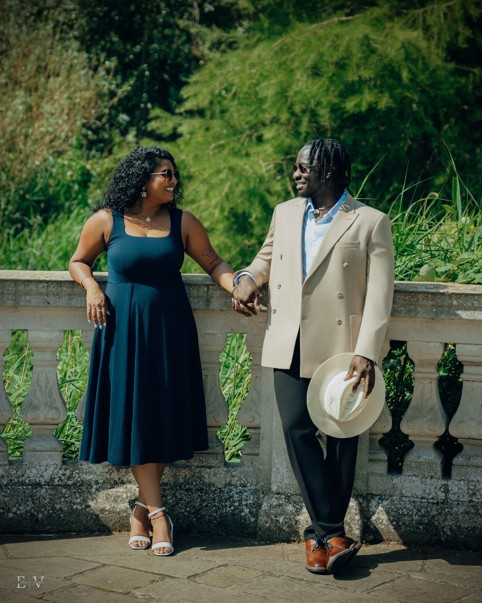 Standing a little apart at a stone balustrade with hands linked, the straw hat held low in one hand.