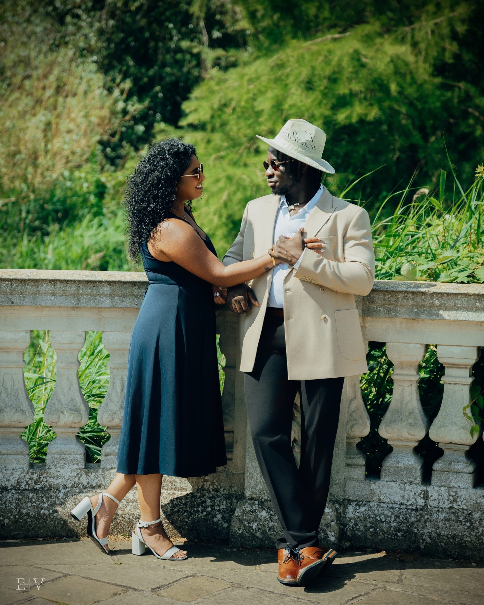 Leaning against the sunlit balustrade in conversation, hands together, hat on.