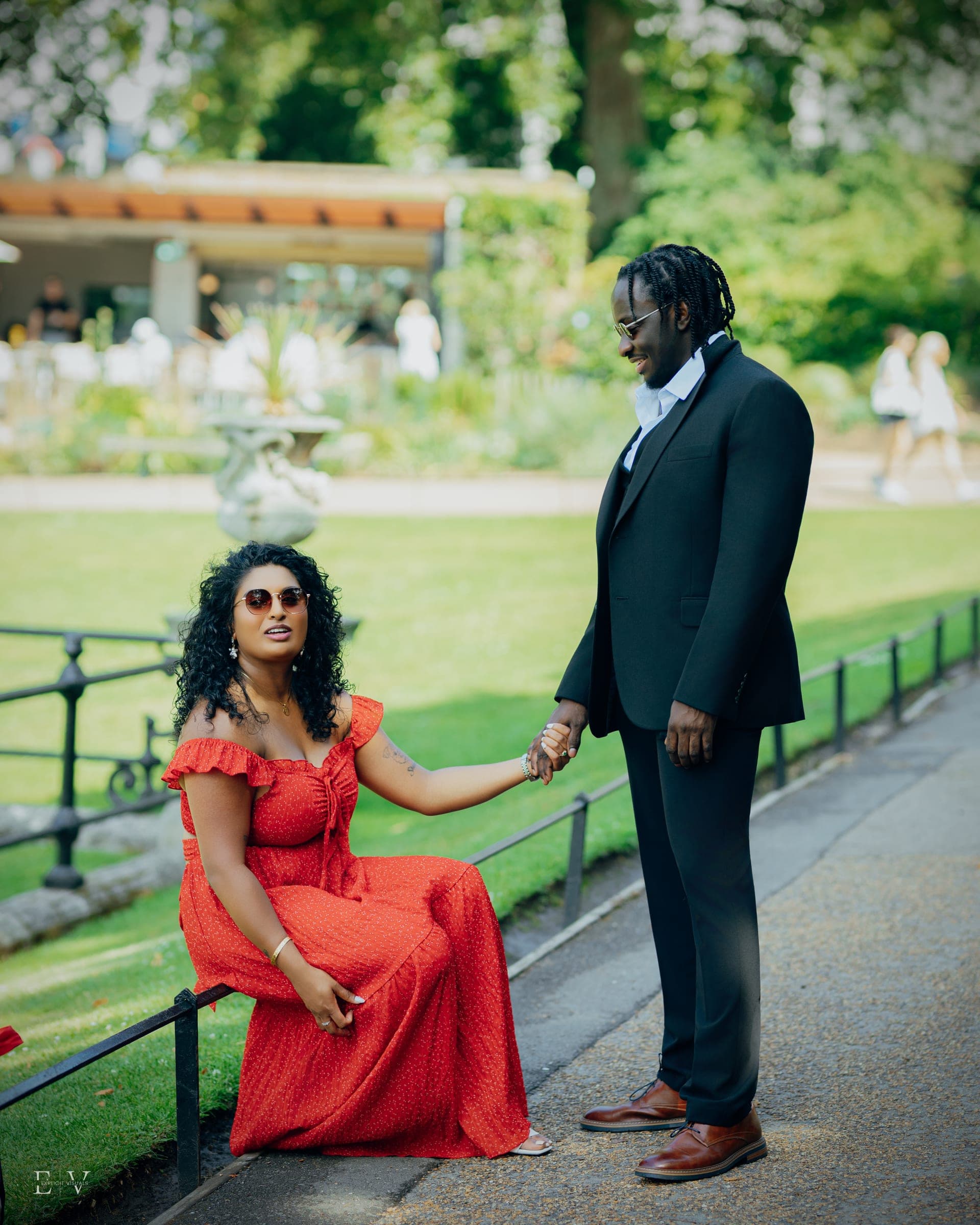 A change of outfit: seated on a low park railing in a long red dress, one hand reaching out to the other, who stands in a black waistcoat and tie.