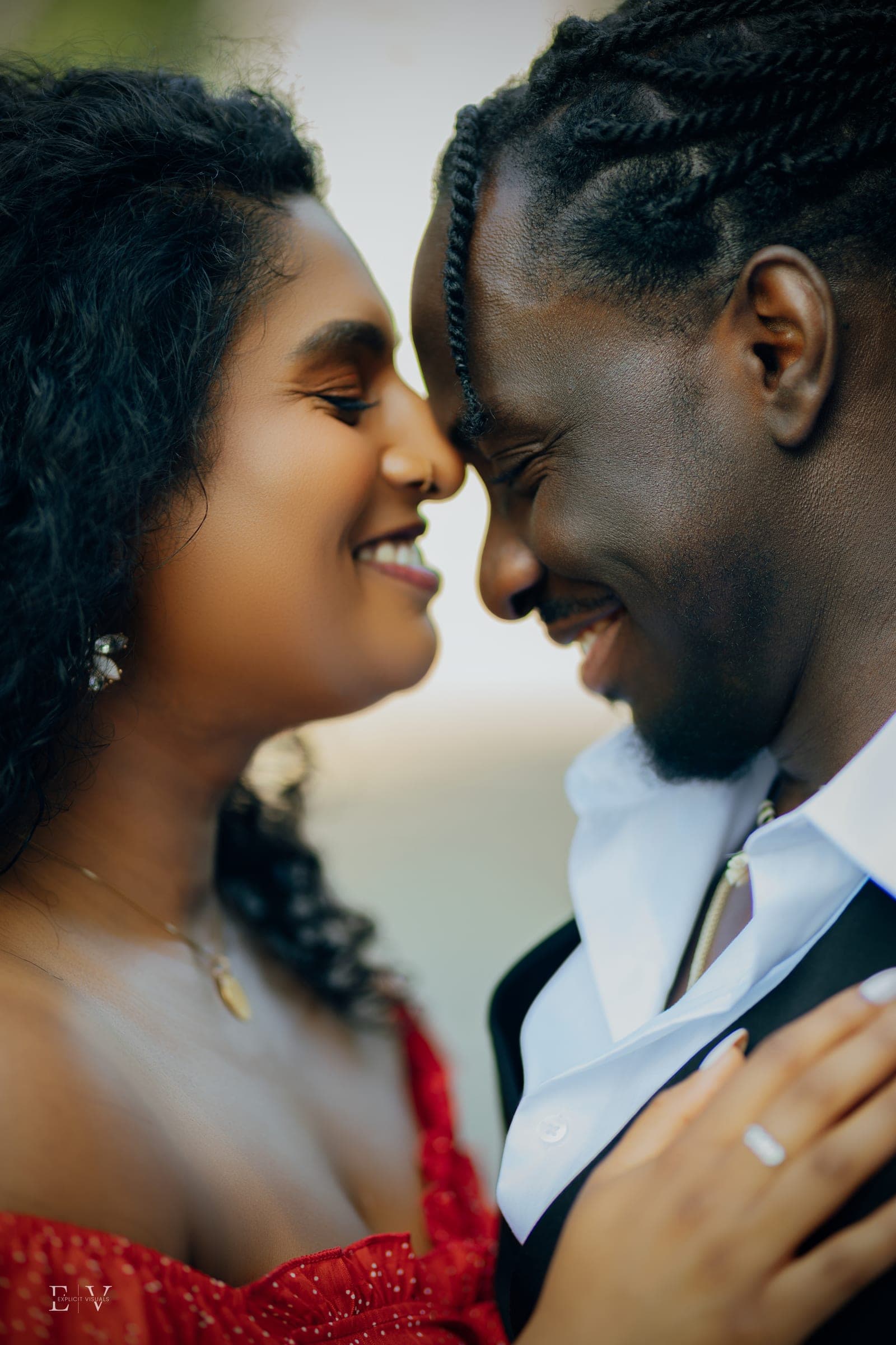 Foreheads together and eyes closed, both smiling; an engagement ring catches the light on the hand resting against a white shirt.