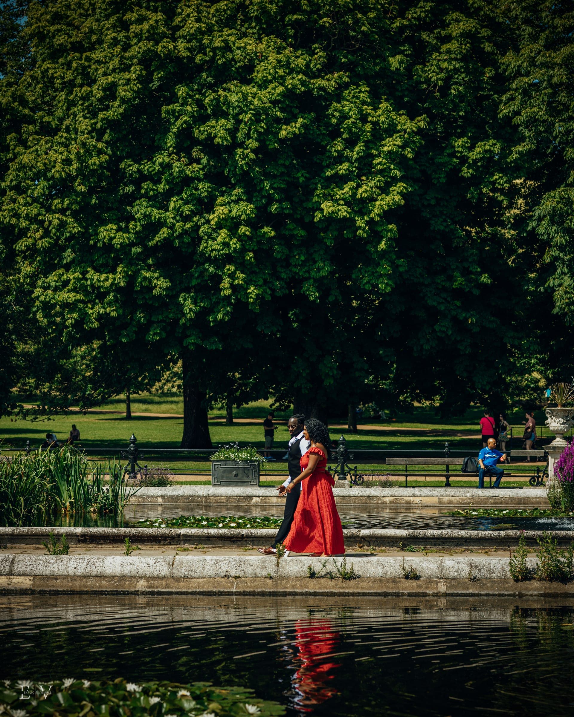 A wide frame of the two small on the path beside a still pool, dwarfed by one huge dark tree, the red dress the only colour in the picture.
