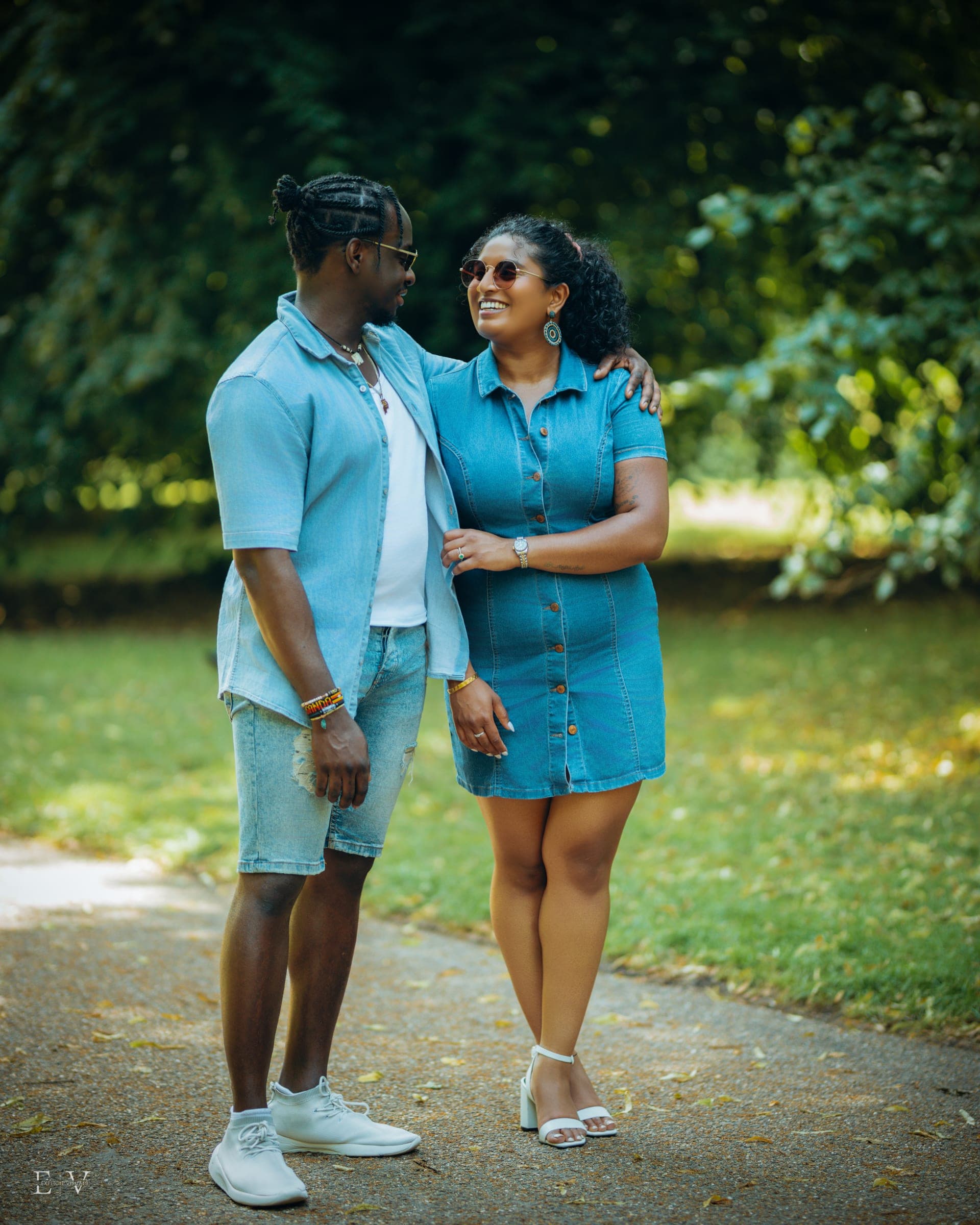 A third look, both in matching pale blue denim, arms around each other on a shaded park path, mid-laugh.