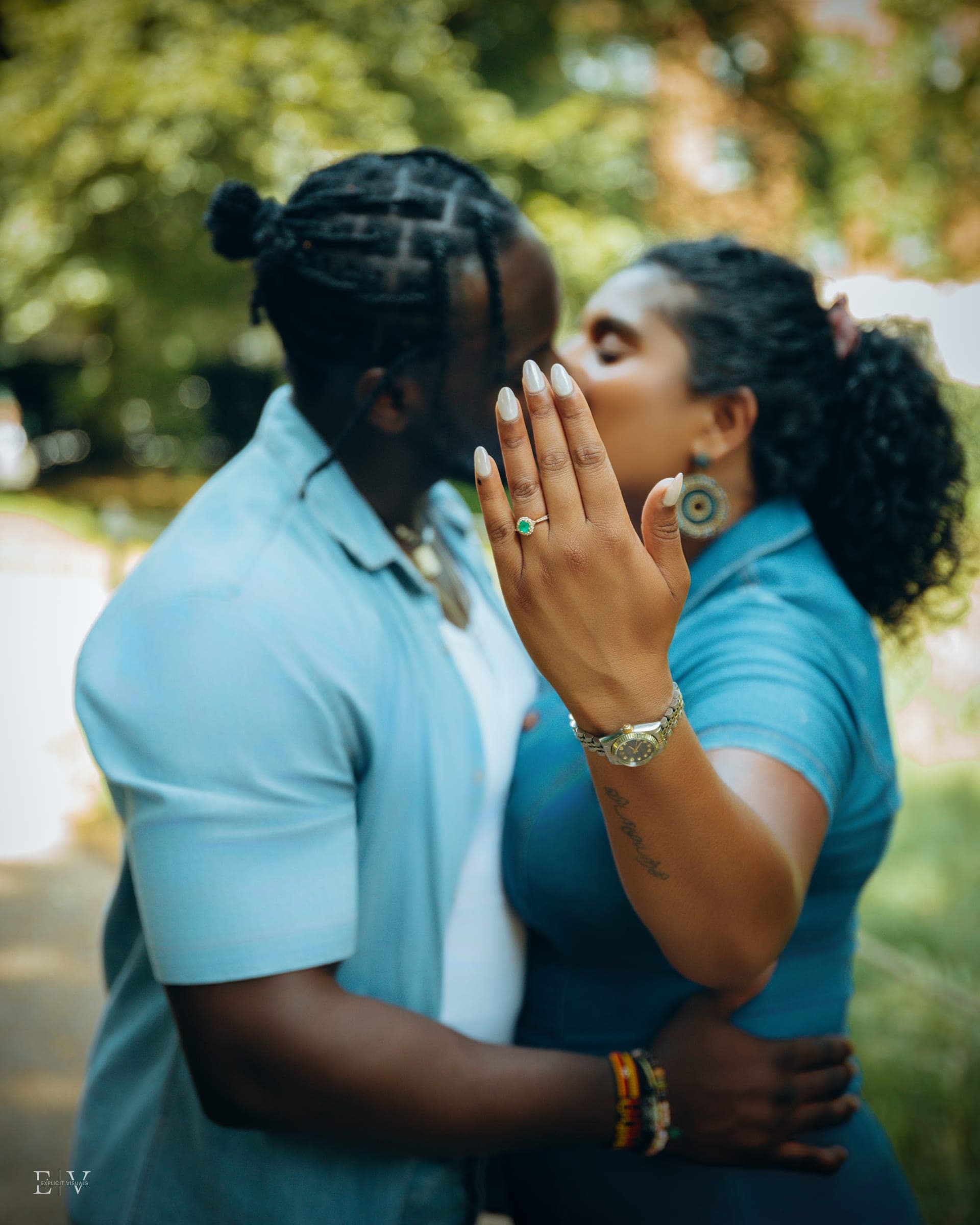 A kiss hidden behind a hand raised to the camera, the engagement ring and a gold watch sharp in the foreground.