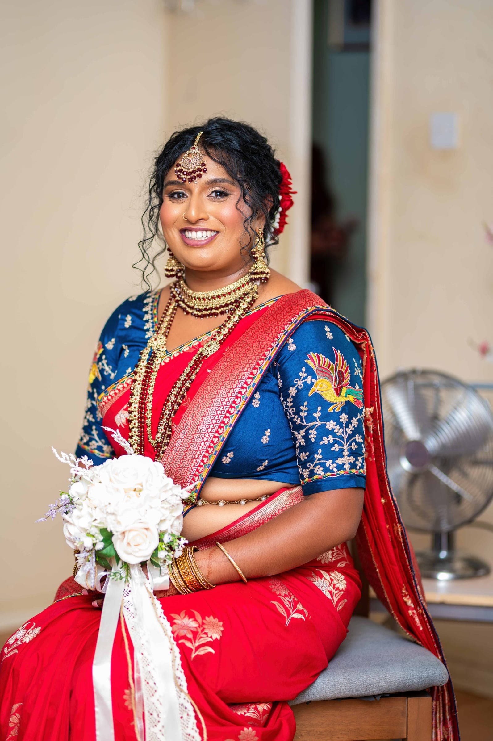 The bride seated at home in a red silk saree and a blue blouse embroidered with a bird, wearing layered gold and garnet jewellery and holding a white rose bouquet.