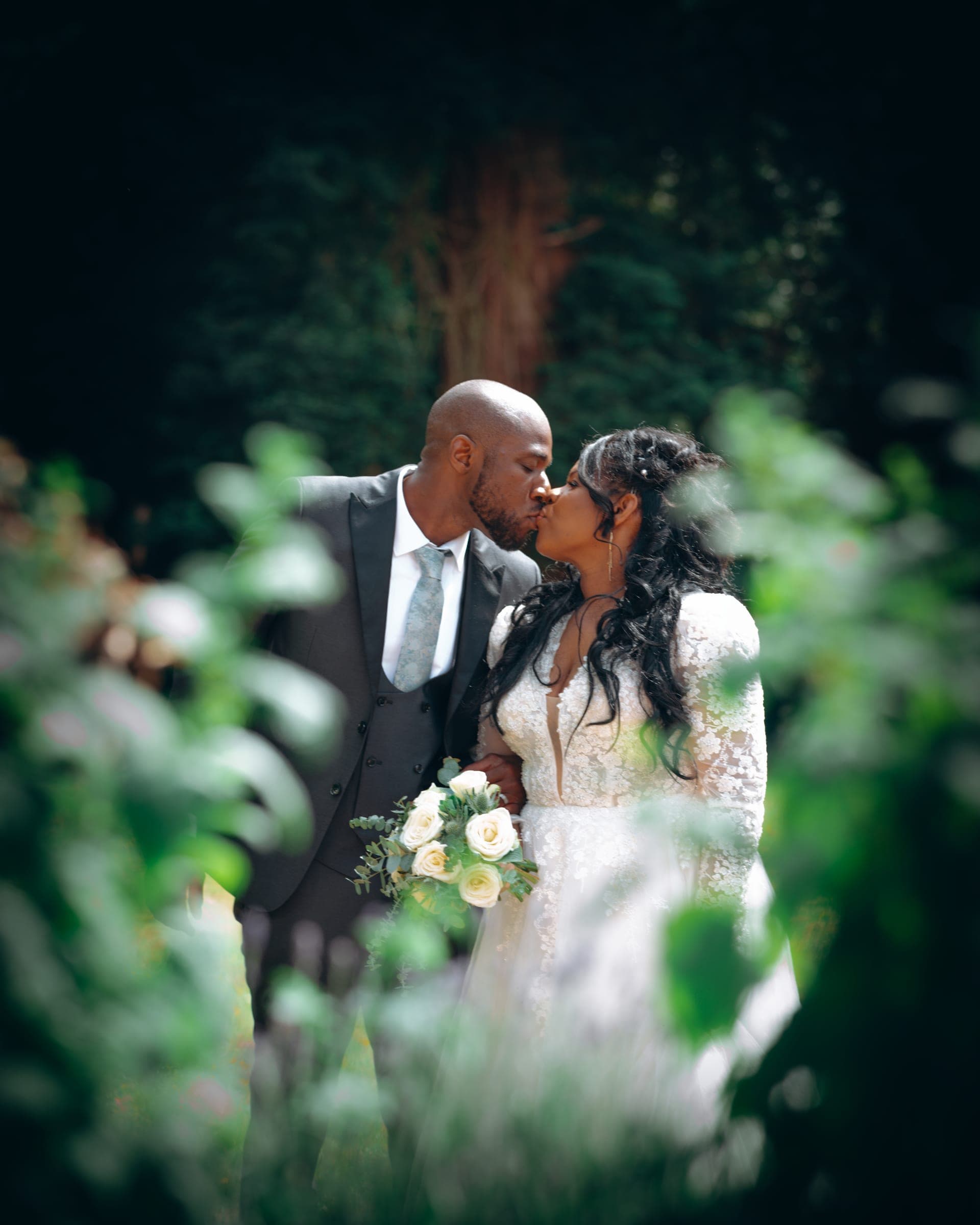A kiss in the garden shot through foreground leaves, the two of them sharp at the centre of a dark green frame.