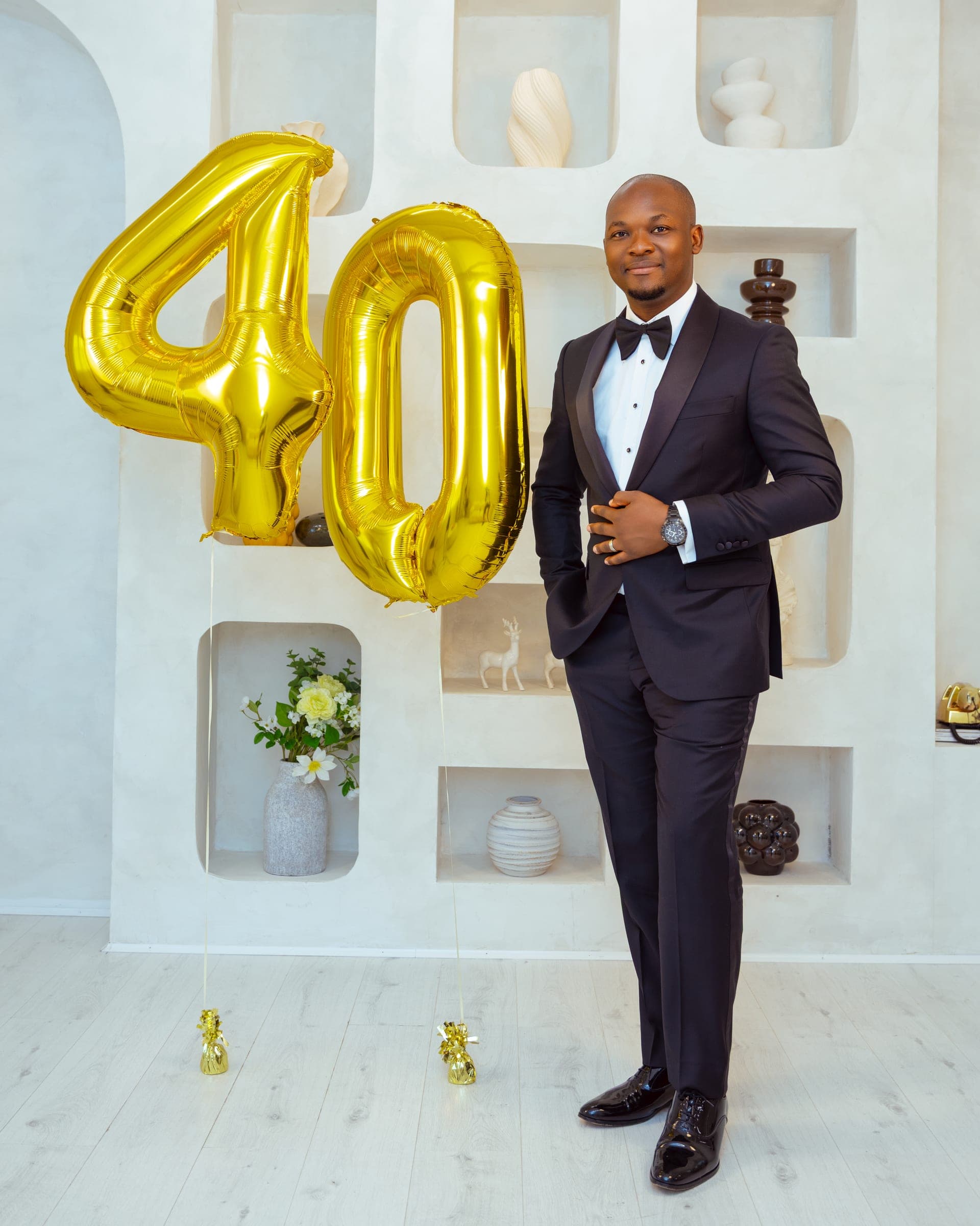 Full length in a black tuxedo and bow tie beside two giant gold foil balloons reading 40, in a white studio set of arched alcoves.
