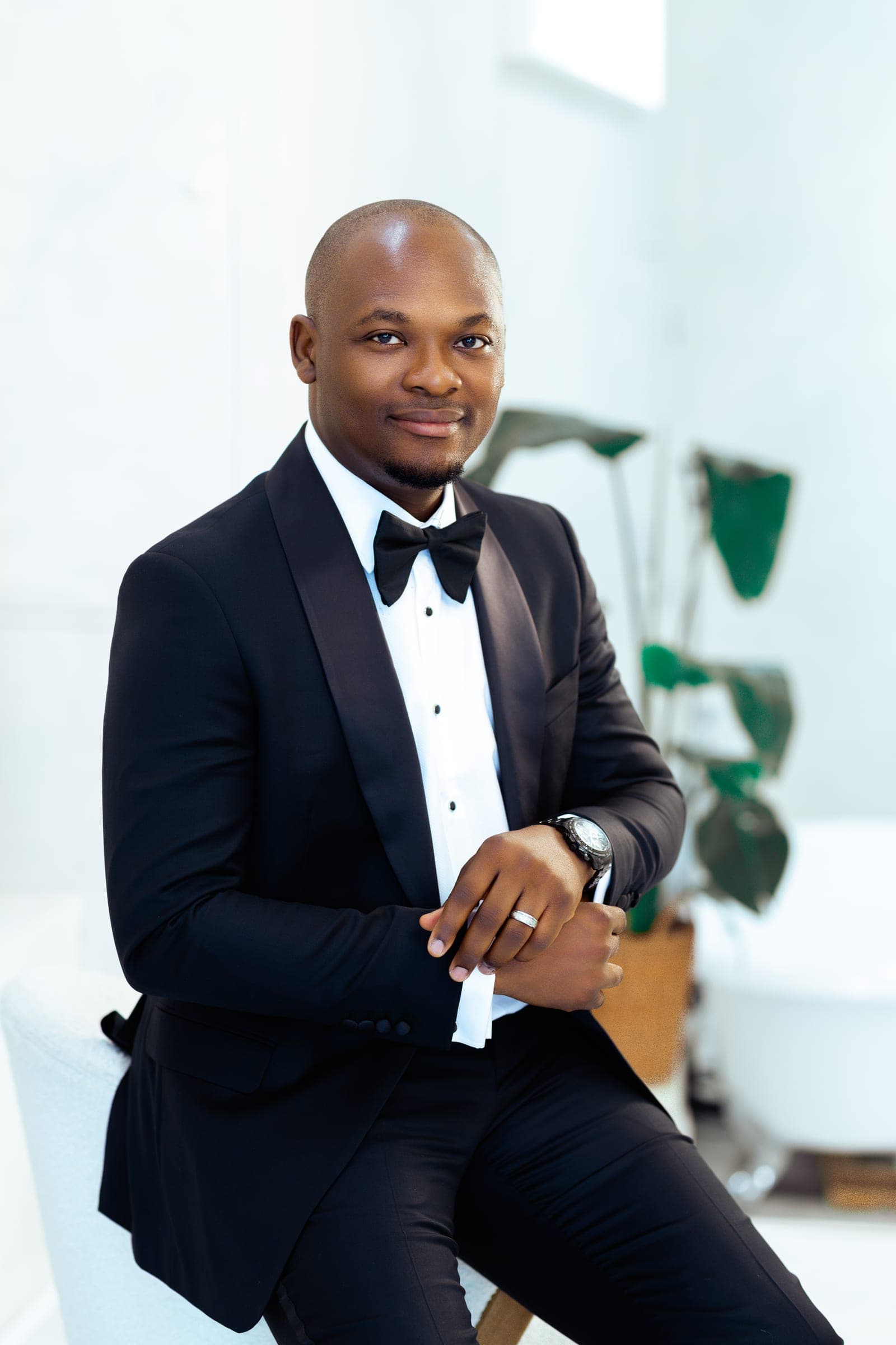A three-quarter portrait in the tuxedo, hands together at his cuff and a steel watch showing, daylight flooding the white studio behind him.
