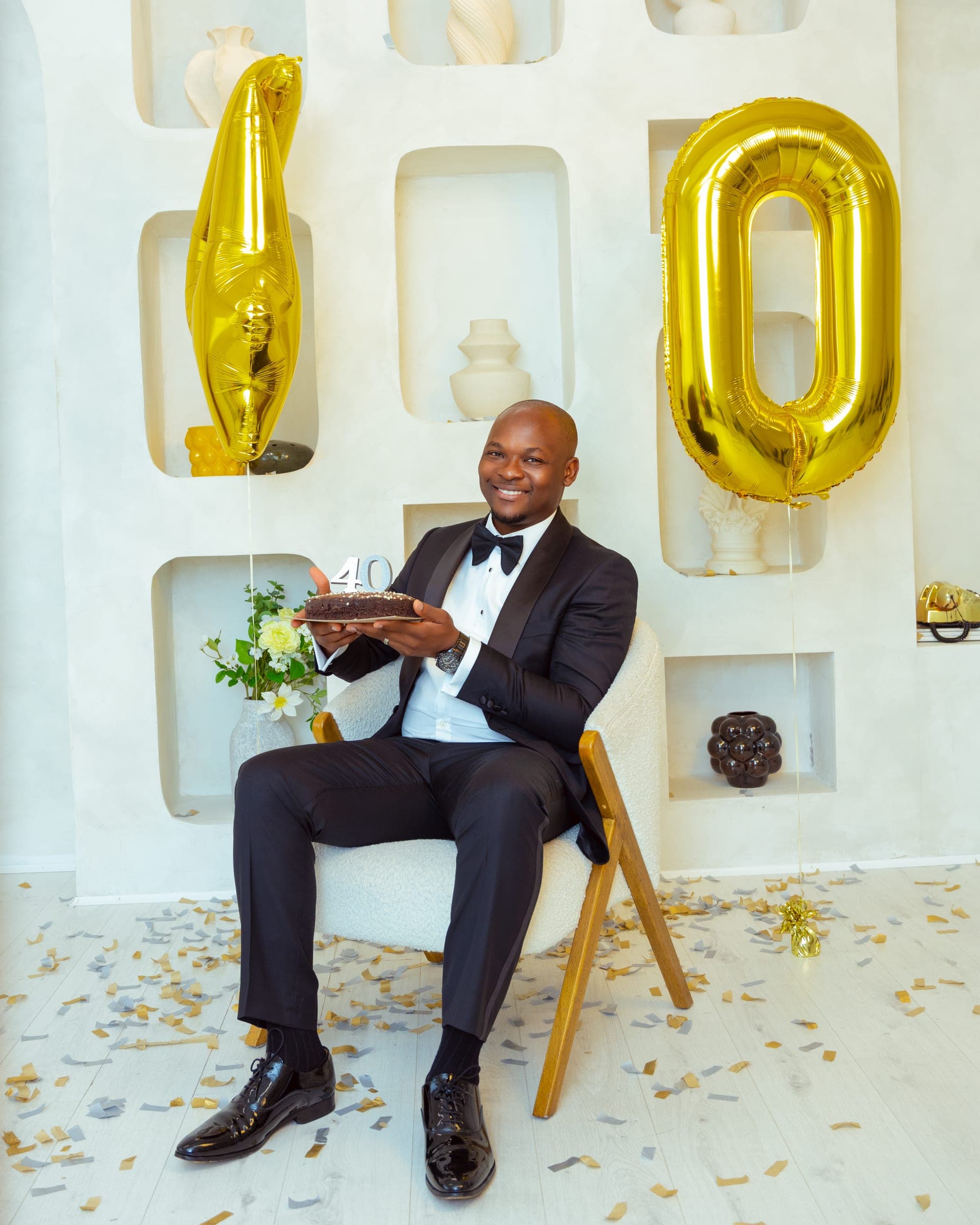 Seated between the gold 4 and 0 balloons holding a small dark birthday cake, gold confetti scattered across the studio floor.