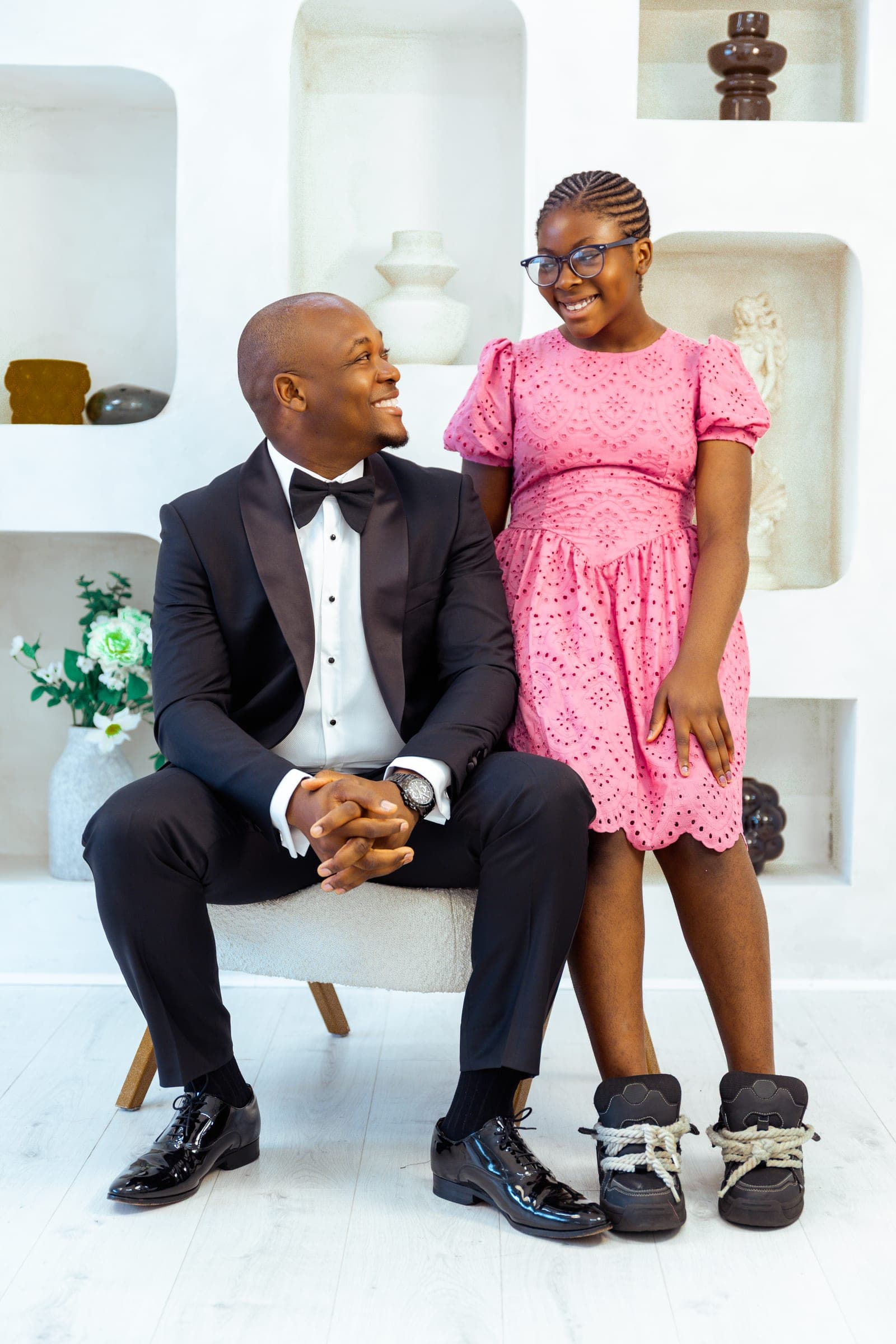 His eldest daughter stands beside the chair in a pink broderie dress and glasses; the two of them look at each other and laugh.