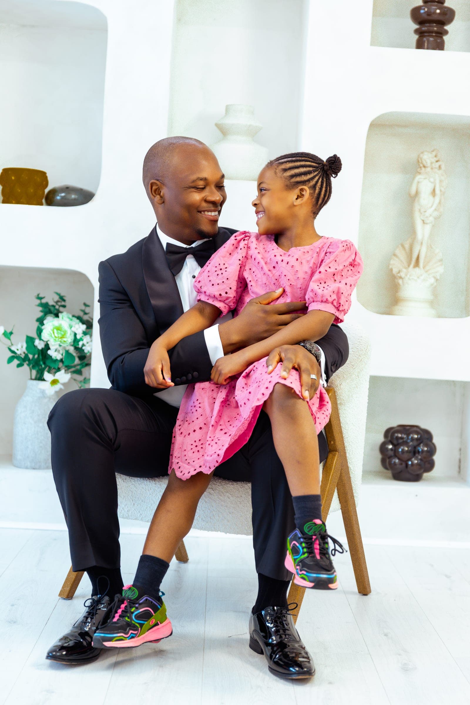 His youngest daughter sits across his lap in a matching pink dress, both mid-laugh, her trainers bright against his black suit.