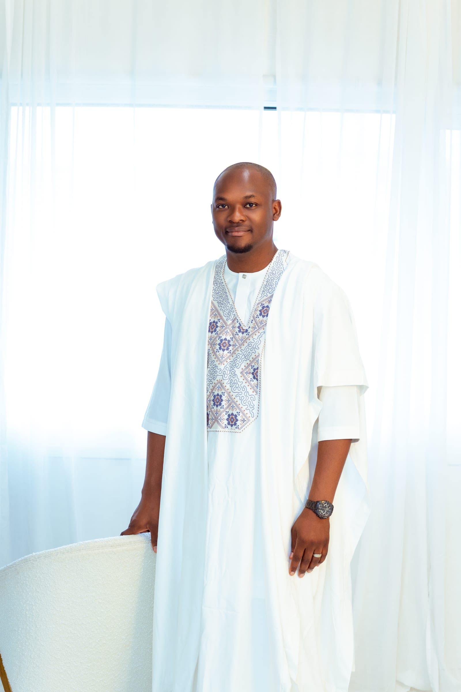 A change into white Nigerian dress — an embroidered agbada and cap — standing against bright curtained windows with a hand on a chair back.
