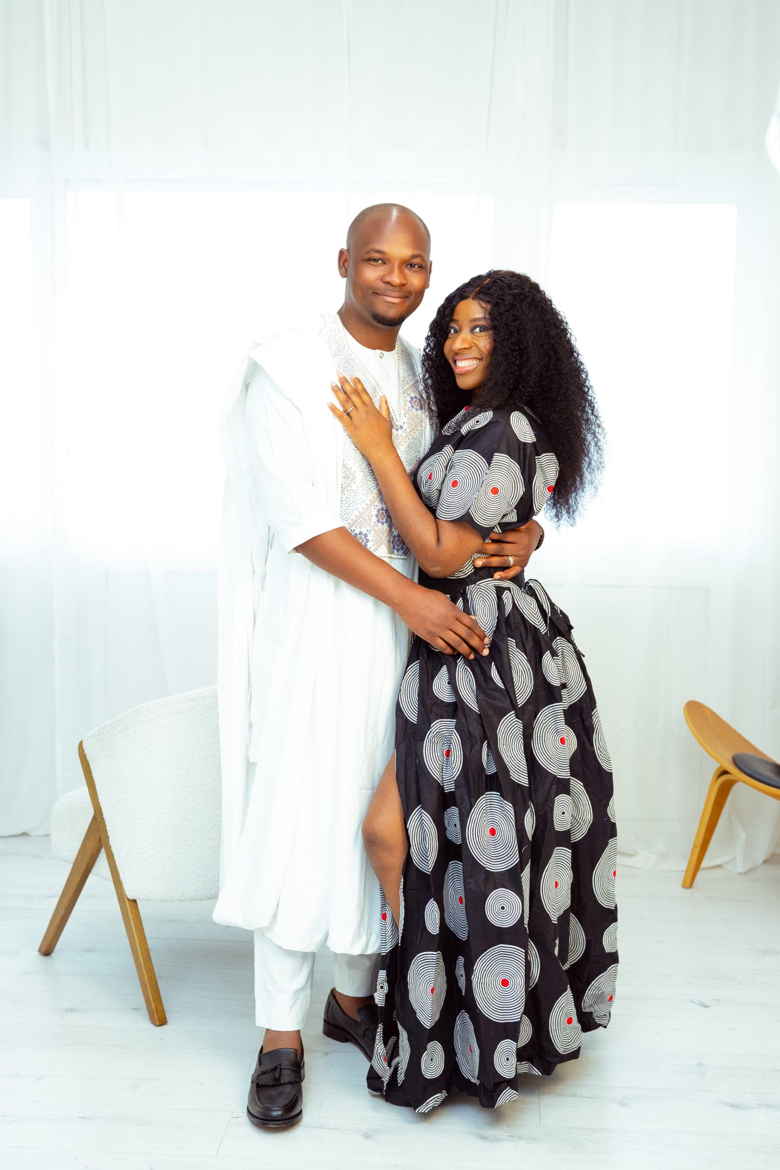 The couple stand together, he in the white agbada and she in a black and white printed dress, arms wrapped around each other.