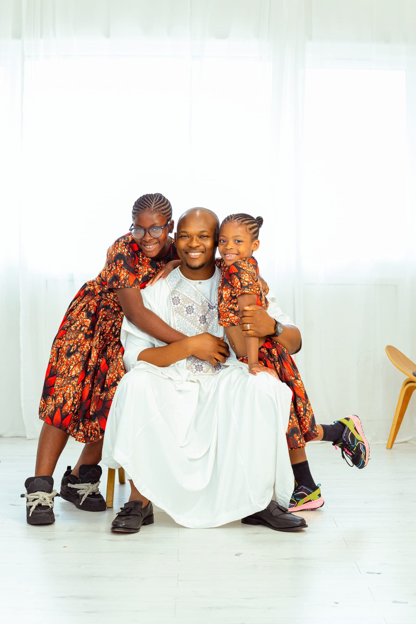 Both daughters lean in from either side, arms around his neck, all three laughing in front of the white curtains.