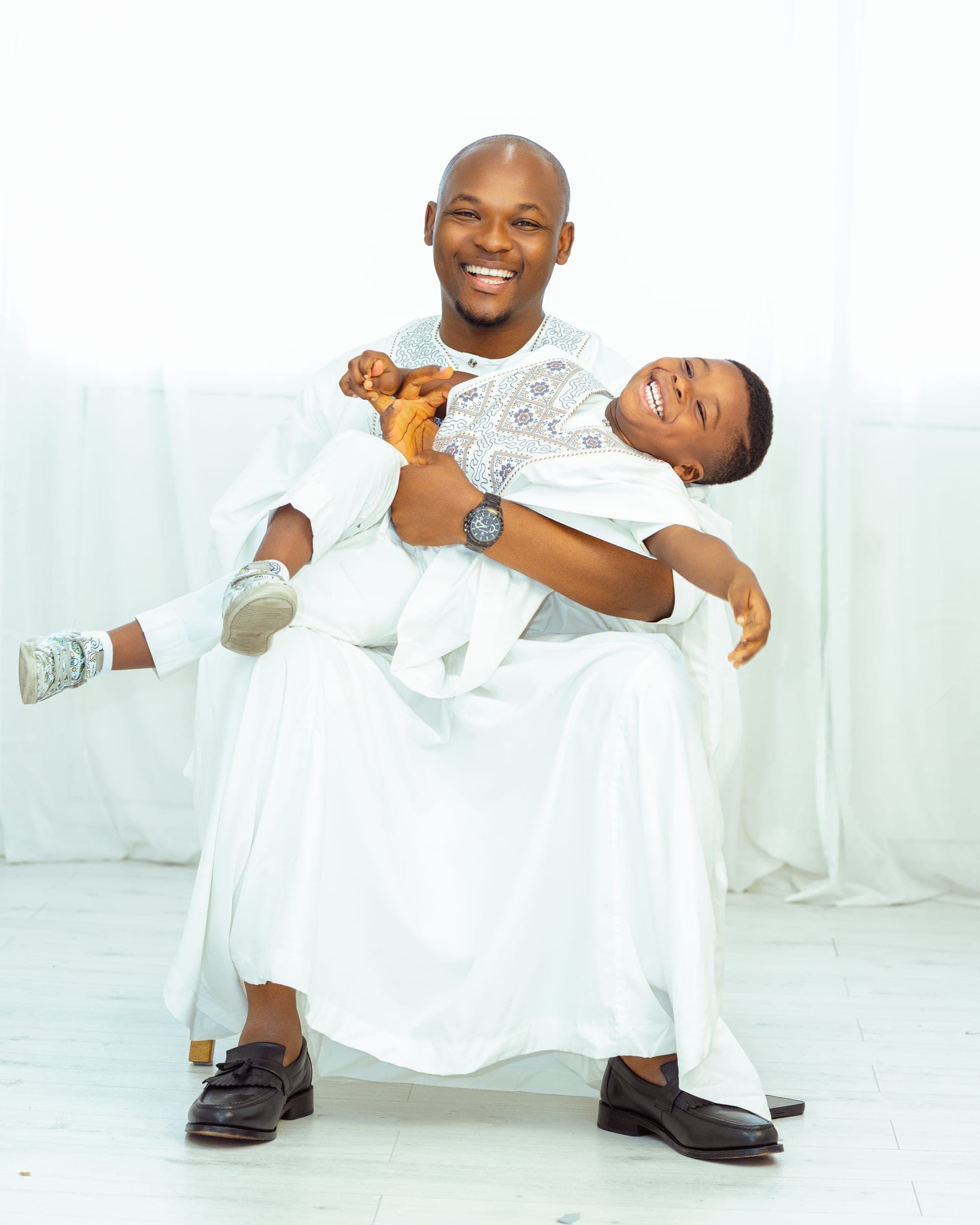 He holds his small son across his lap, the boy laughing with his head thrown back, both in white.