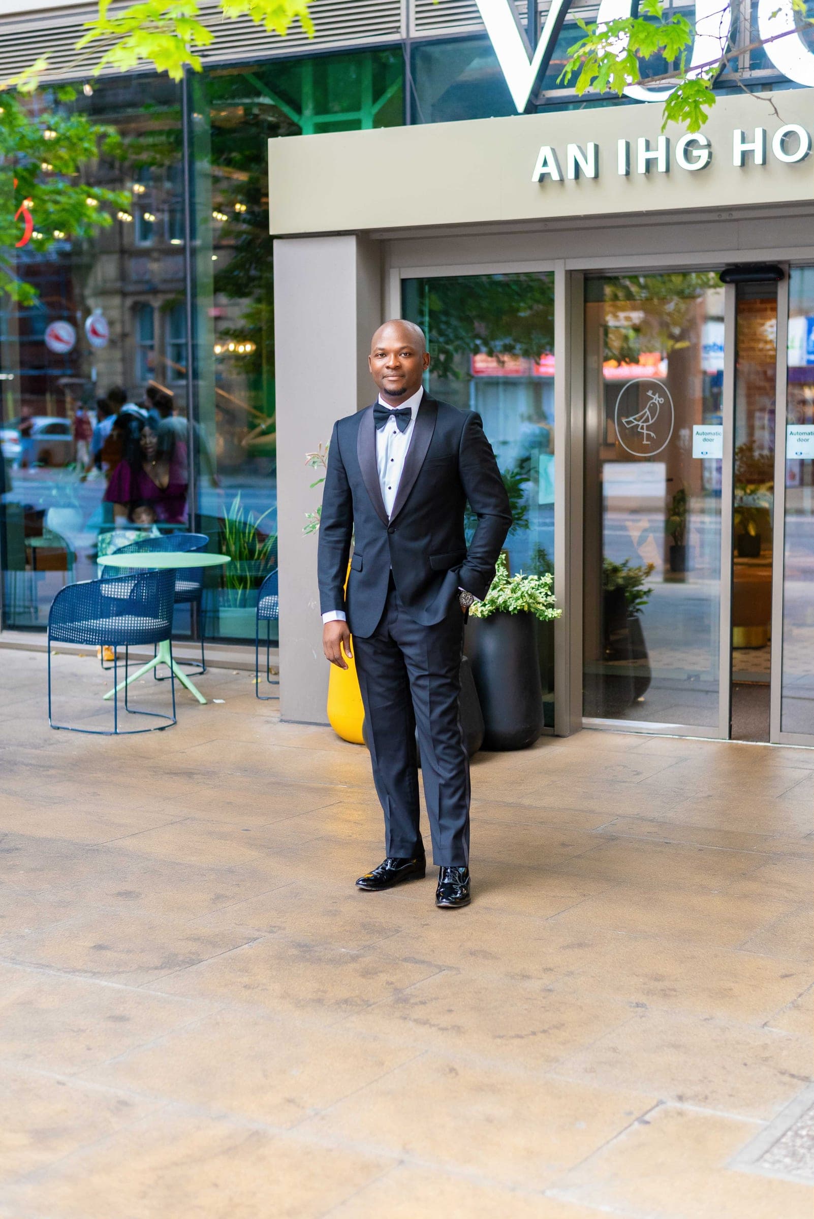 Evening, and he stands in a midnight dinner suit outside the glass doors of a city hotel, a yellow planter behind him.