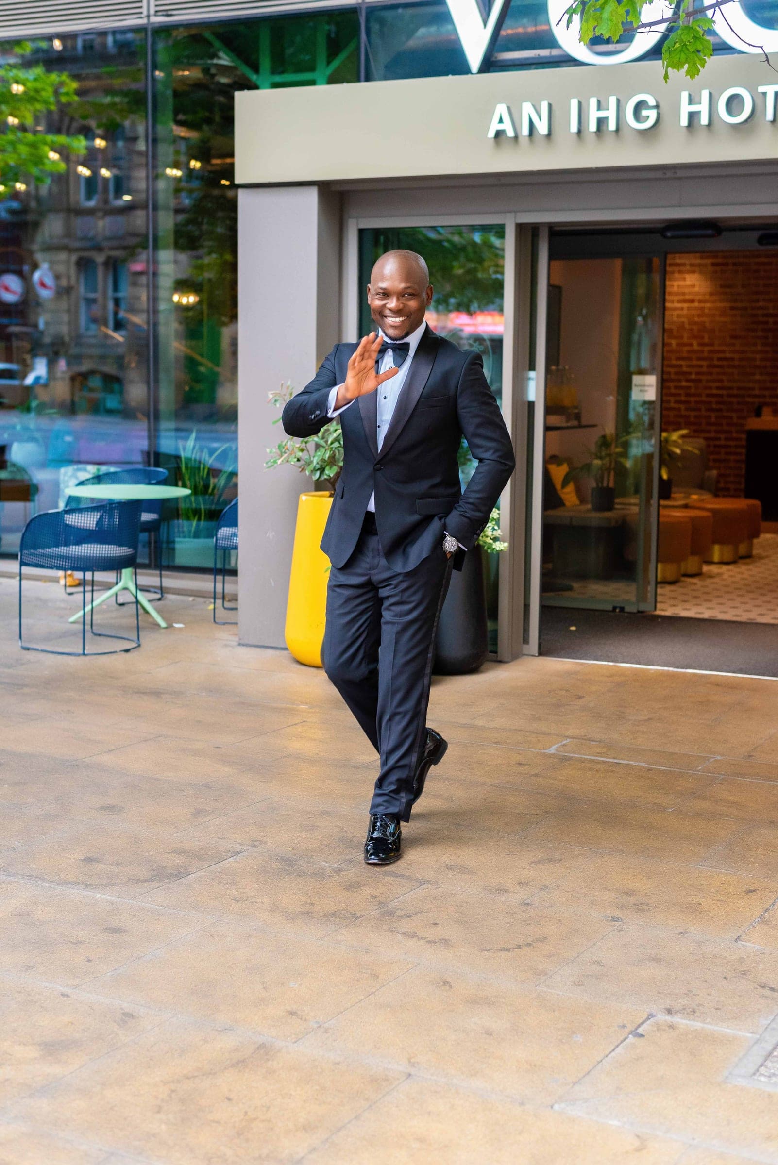 Walking out across the paving with a wave and a wide smile, the hotel entrance open behind him.