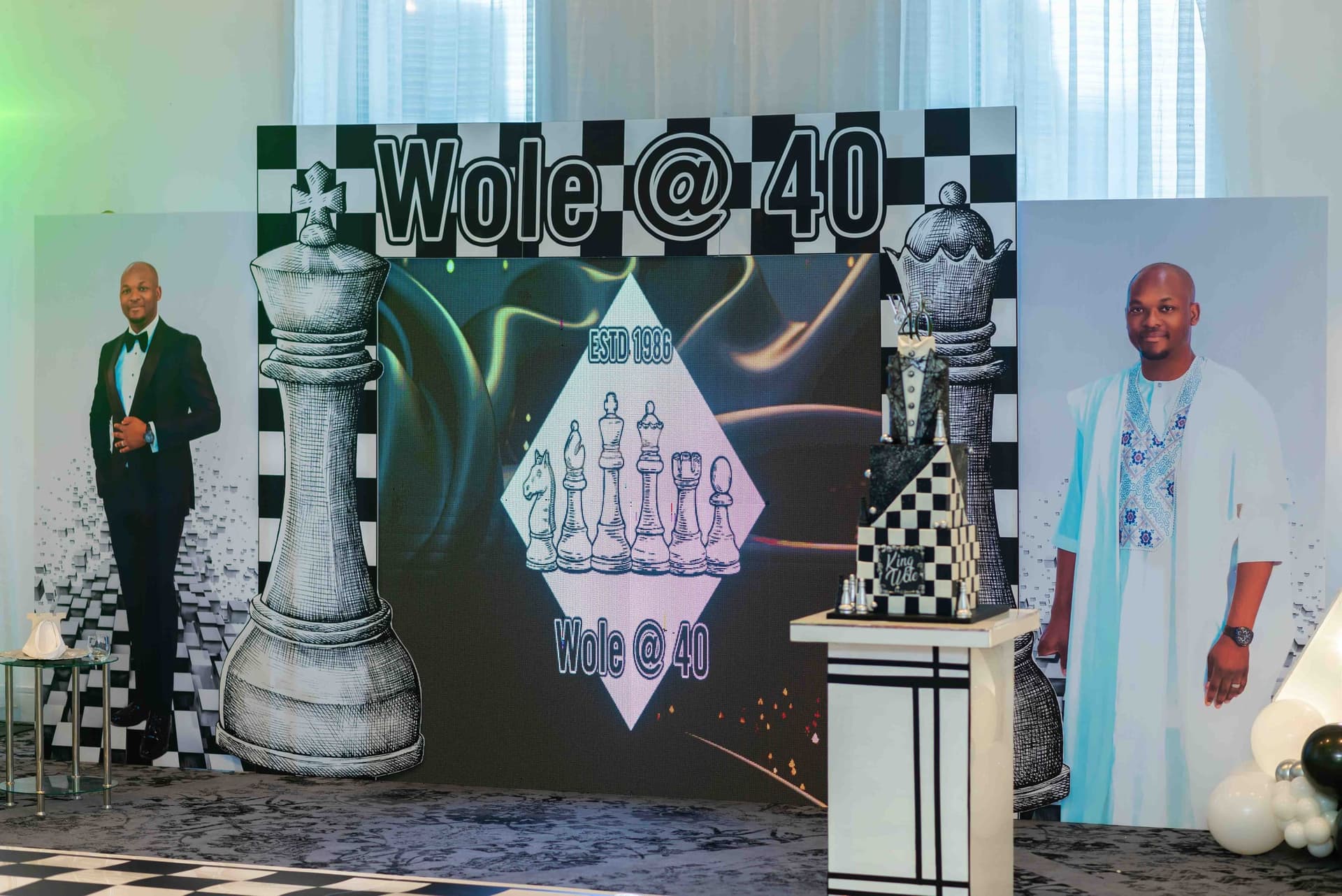 The event backdrop — a chequerboard wall reading Wole @ 40 between two oversized drawn chess pieces, flanked by life-size cutouts of him in black tie and in blue Nigerian dress.