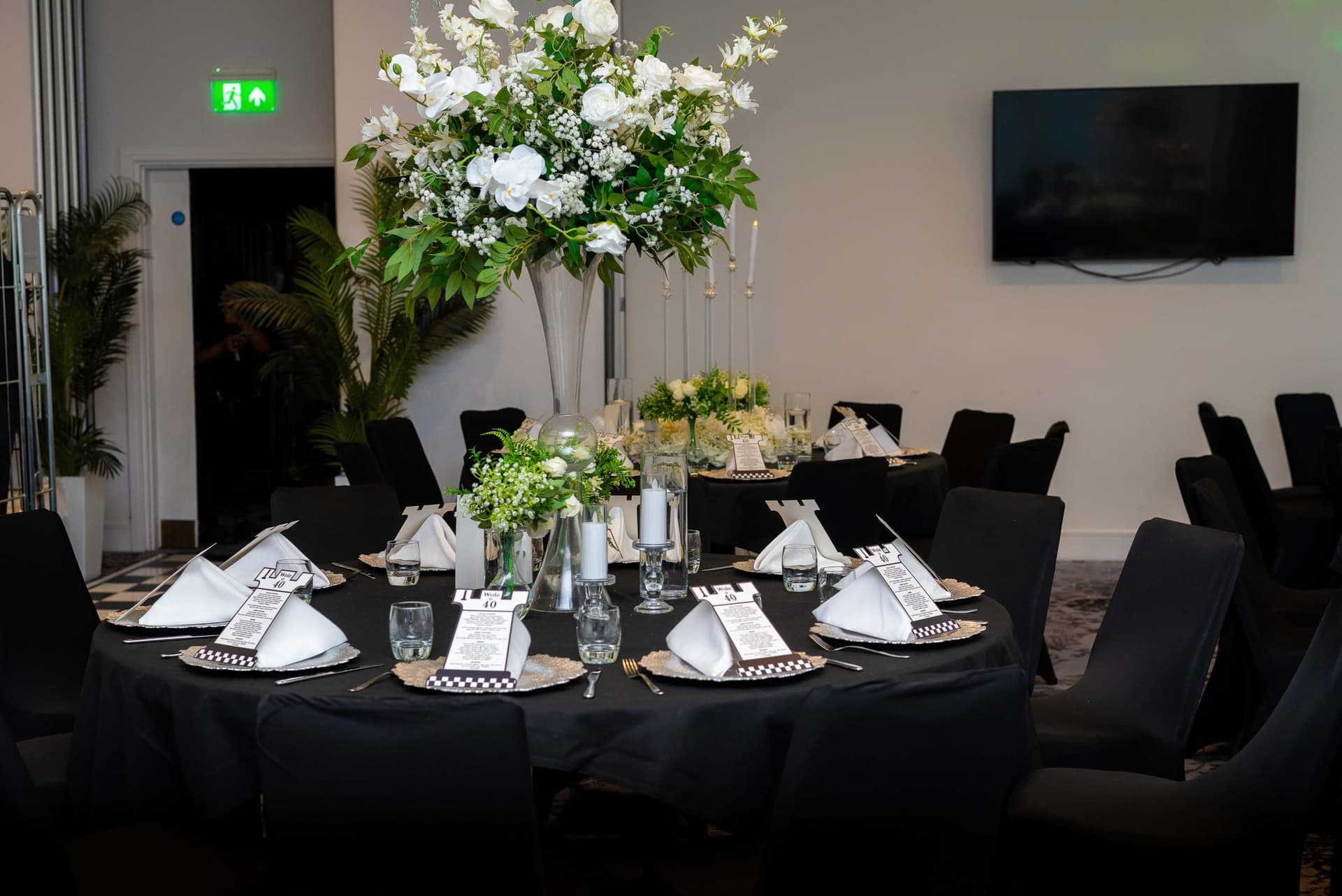A round table laid on black linen with a tall white rose and foliage centrepiece, black chair covers all the way round.