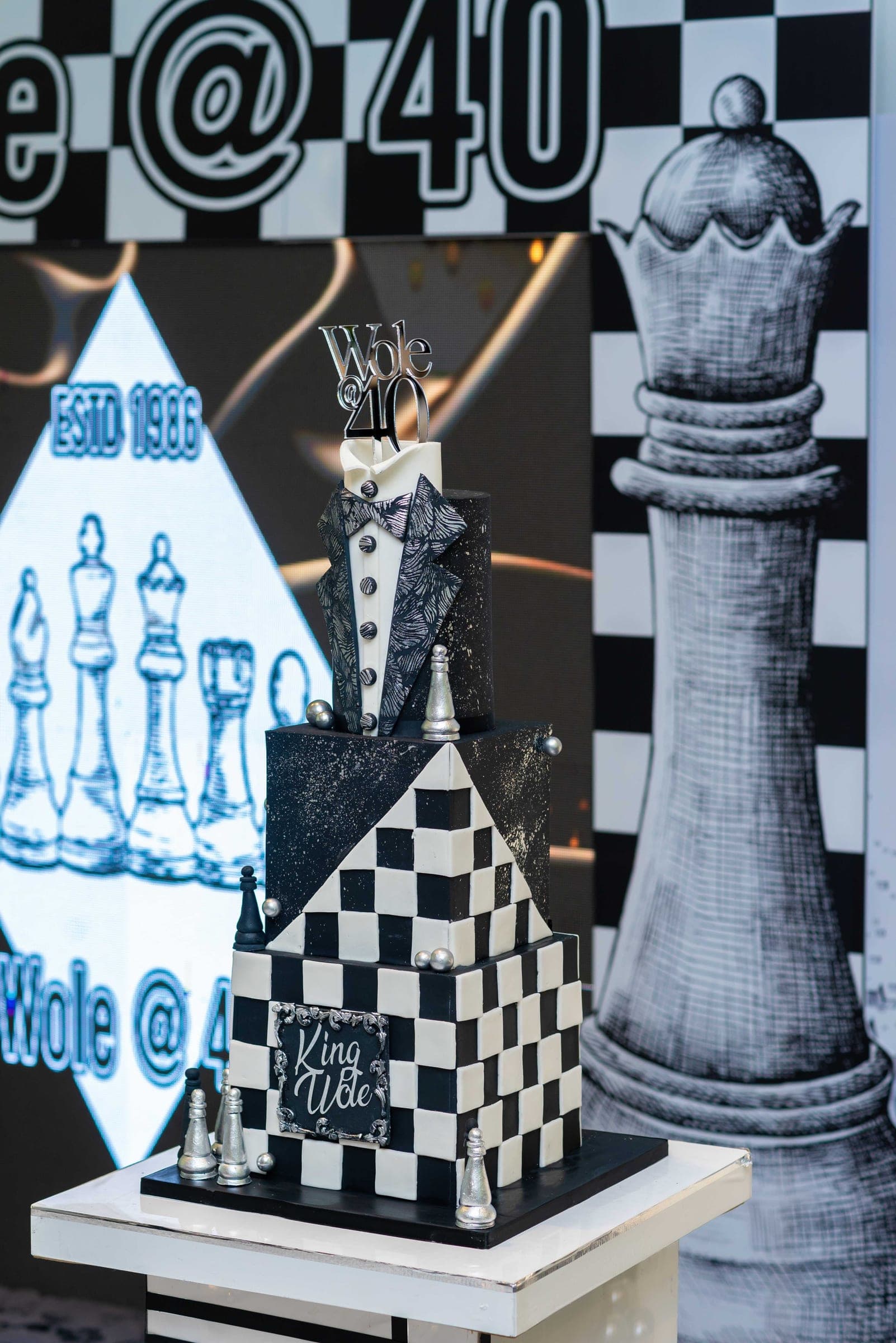 The cake — three tiers in black and white chequerboard, a tuxedo modelled on the top tier and King Wole lettered on the front, silver chess pieces set around the base.