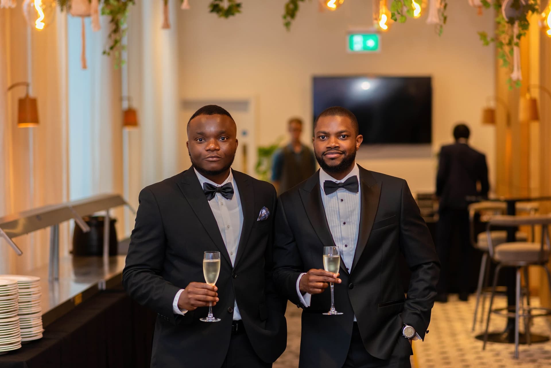 Two guests in dinner jackets stand together in the bar with champagne flutes, warm pendant lights along the wall behind them.