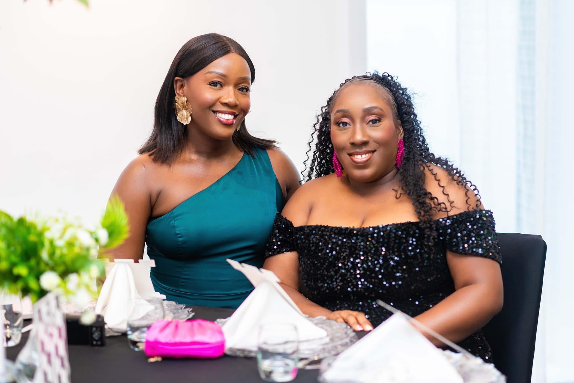 Two guests at their table, one in a teal one-shoulder dress and one in black sequins, both turning to the camera.