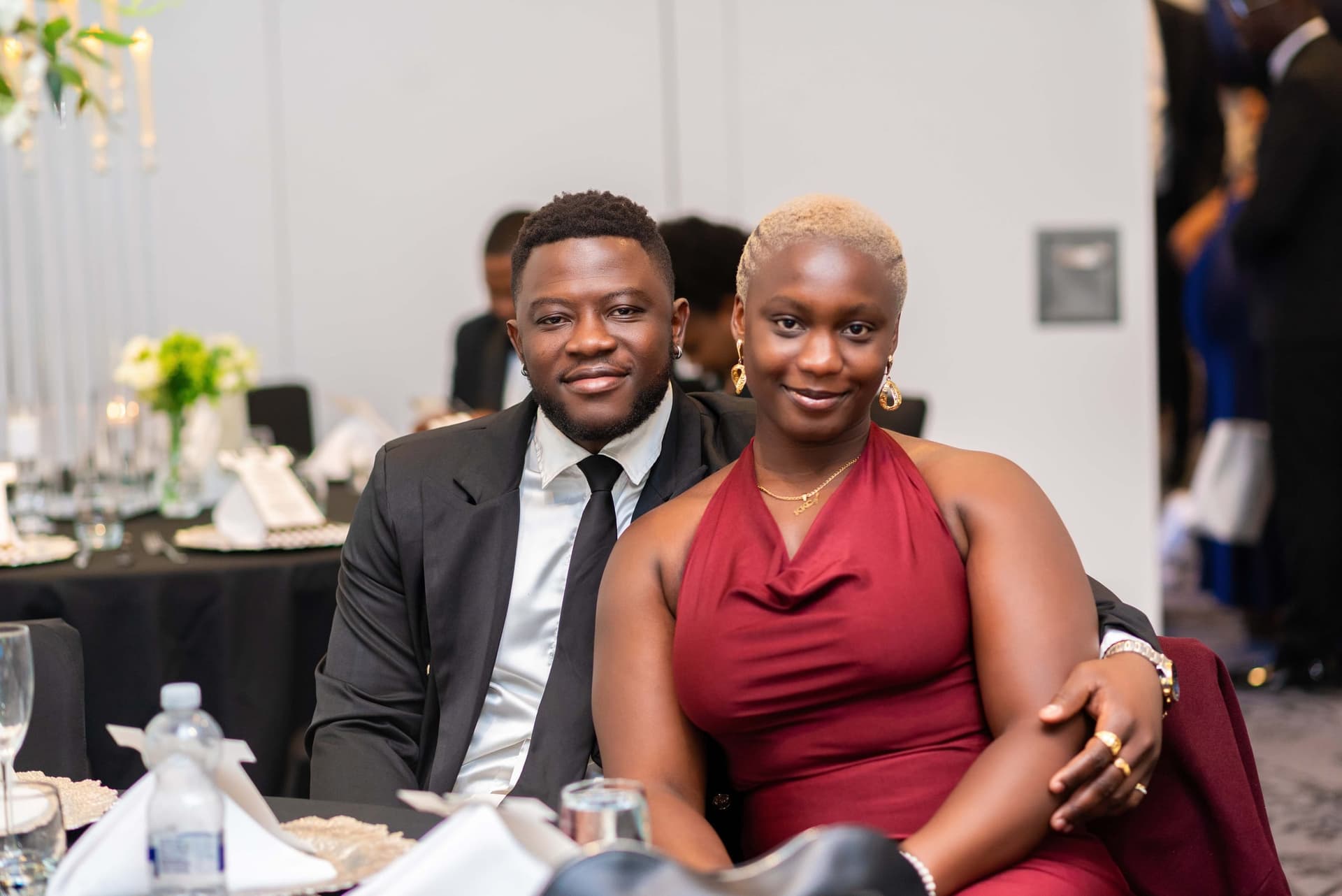 A couple seated at dinner, he in a dinner jacket and she in a red halter dress, his arm resting behind her.