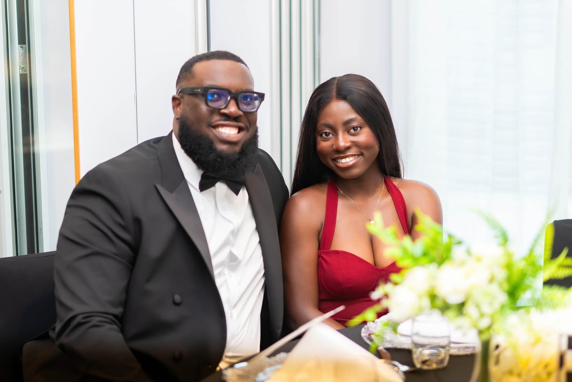 Another couple at the table, he in a black bow tie and she in deep red, a low arrangement of flowers in the foreground.