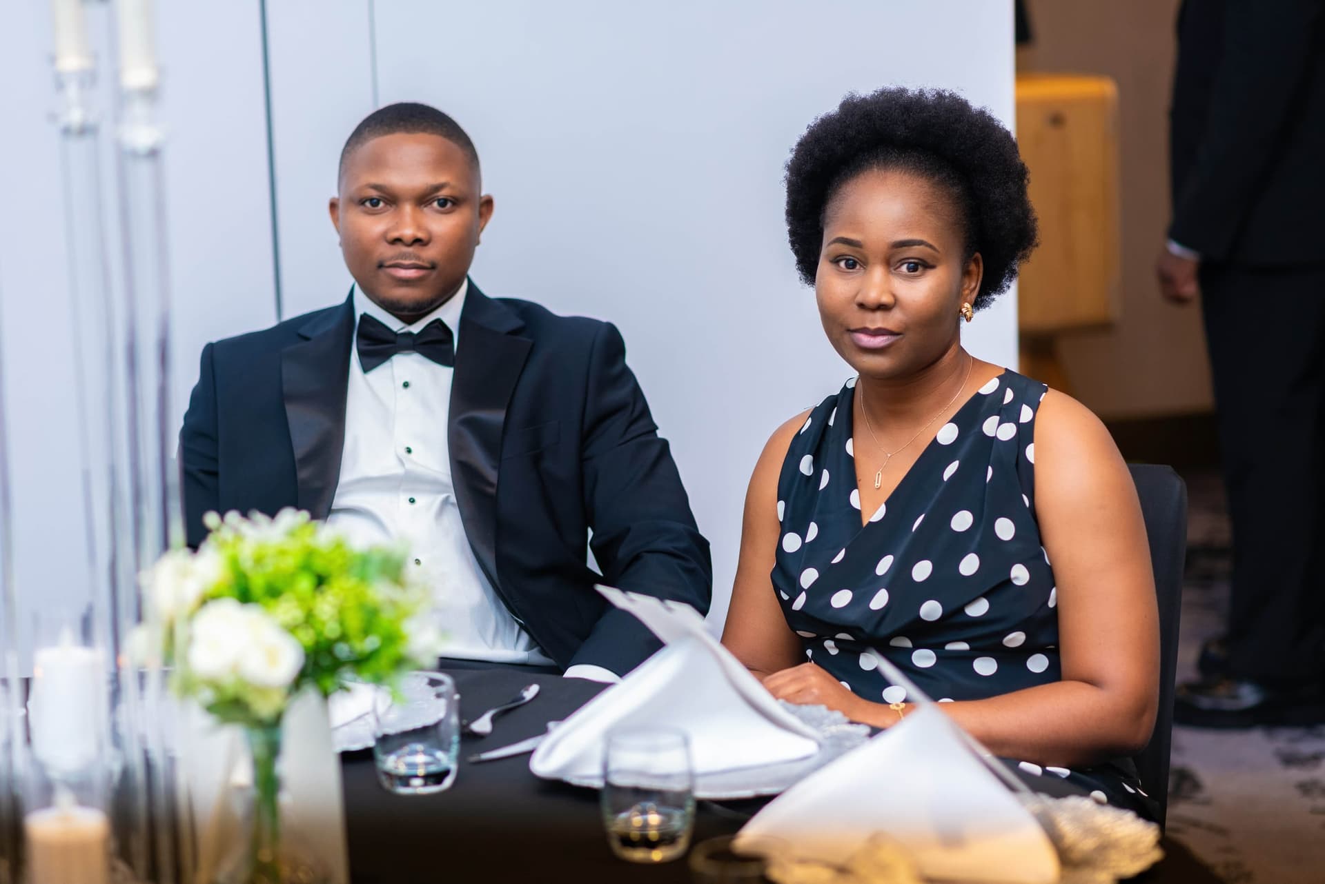 A couple sit quietly at their table, he in black tie, she in a navy polka dot dress, waiting for the speeches to start.