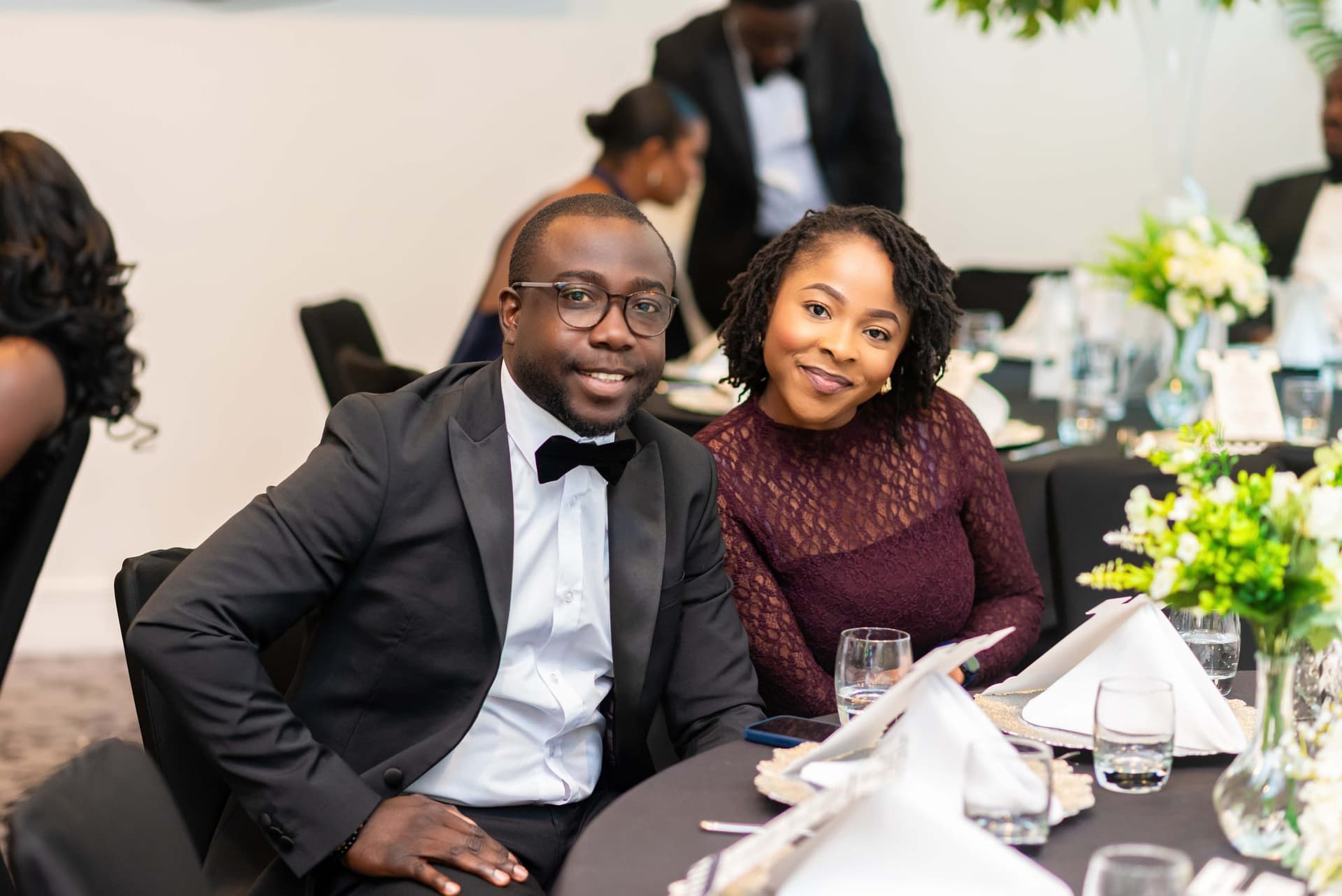 A couple at a round table, he in a dinner jacket and she in burgundy lace, green and white flowers between the place settings.