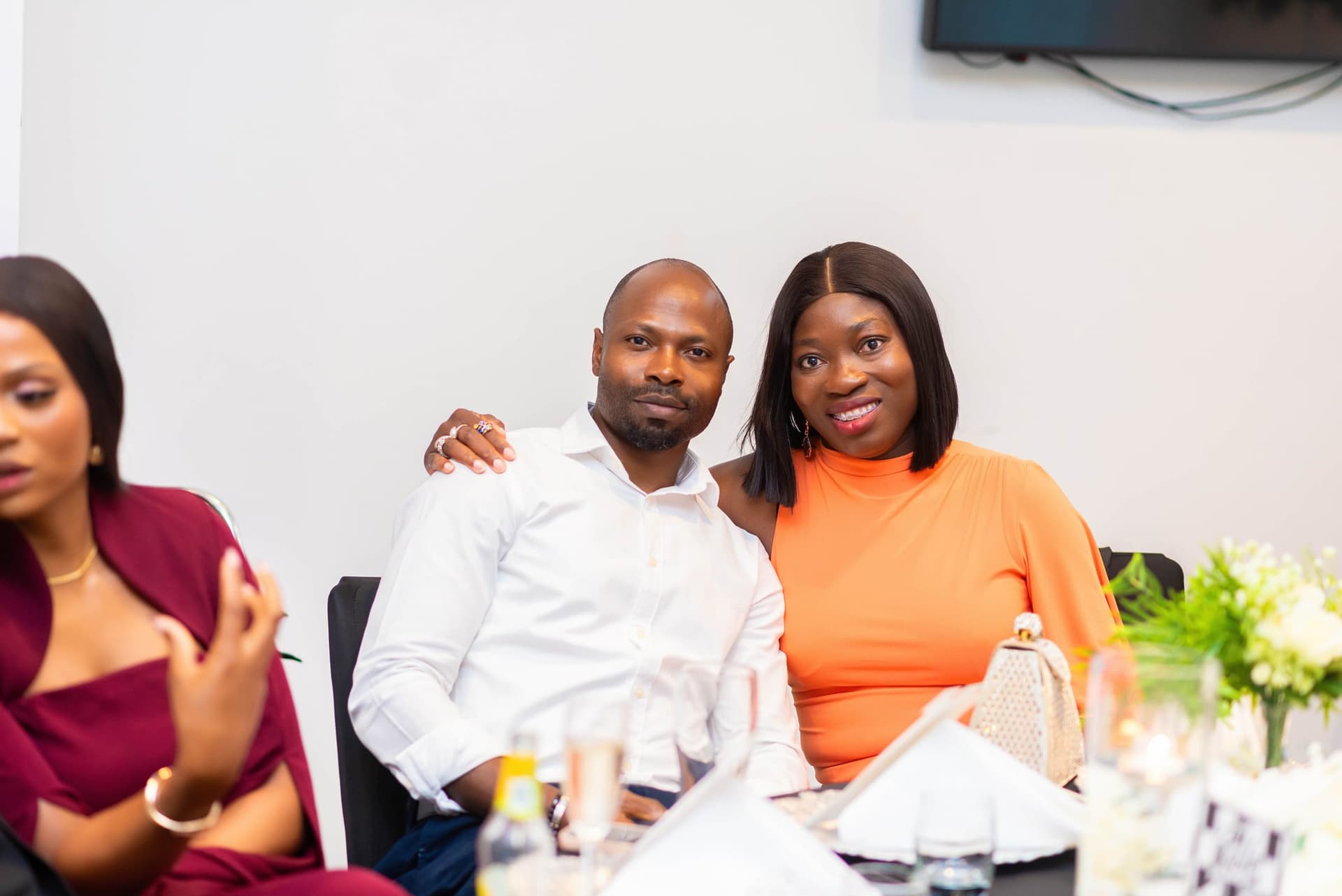 A guest in orange leans into her partner with his arm around her shoulder, the table crowded with glasses in front of them.