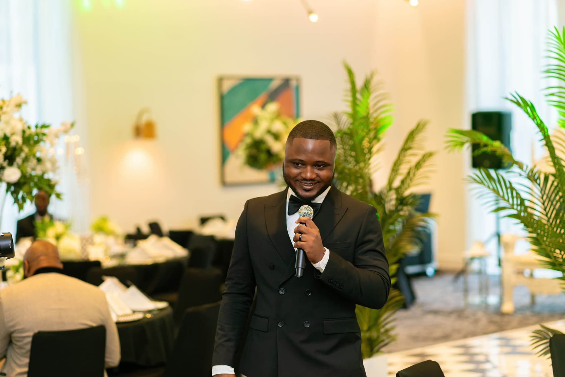 The host stands with a microphone in a double-breasted dinner jacket, opening the evening, palms and set tables behind him.
