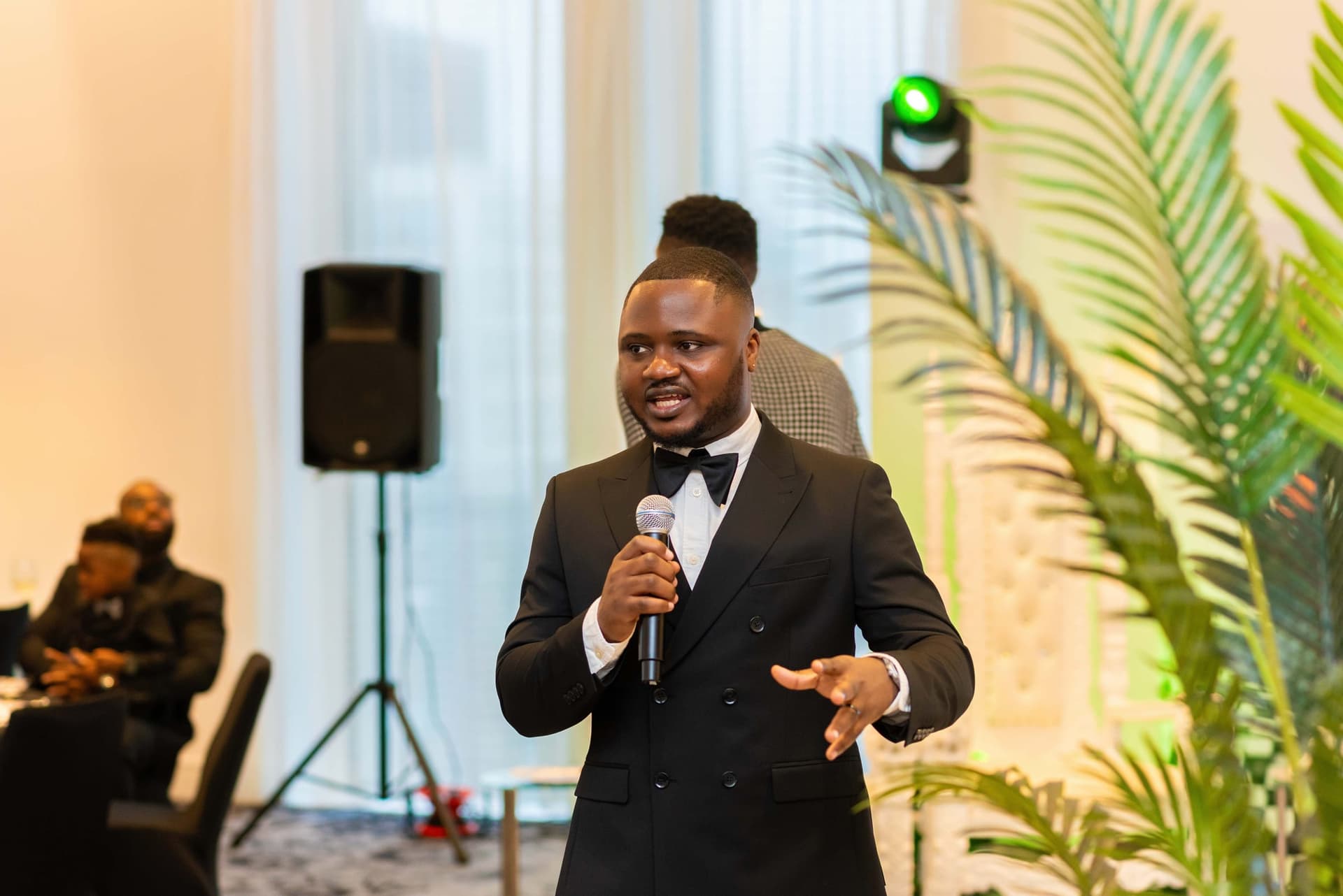 A guest gives a speech from the floor, one hand raised mid-sentence, a stage light throwing green across the wall.
