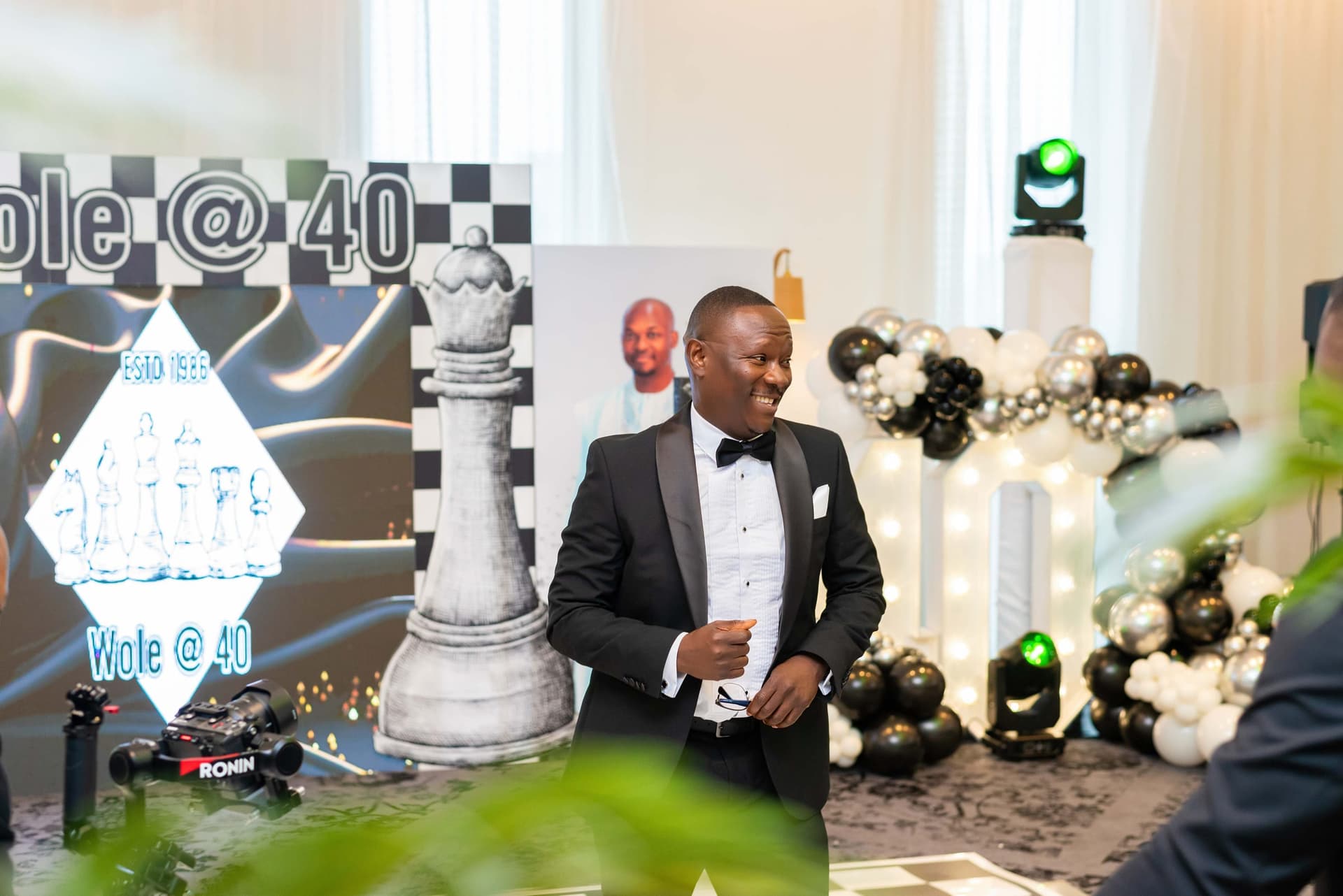 Wole walks in past the chess backdrop in his dinner jacket, laughing, a gimbal-mounted camera just in frame at the left.