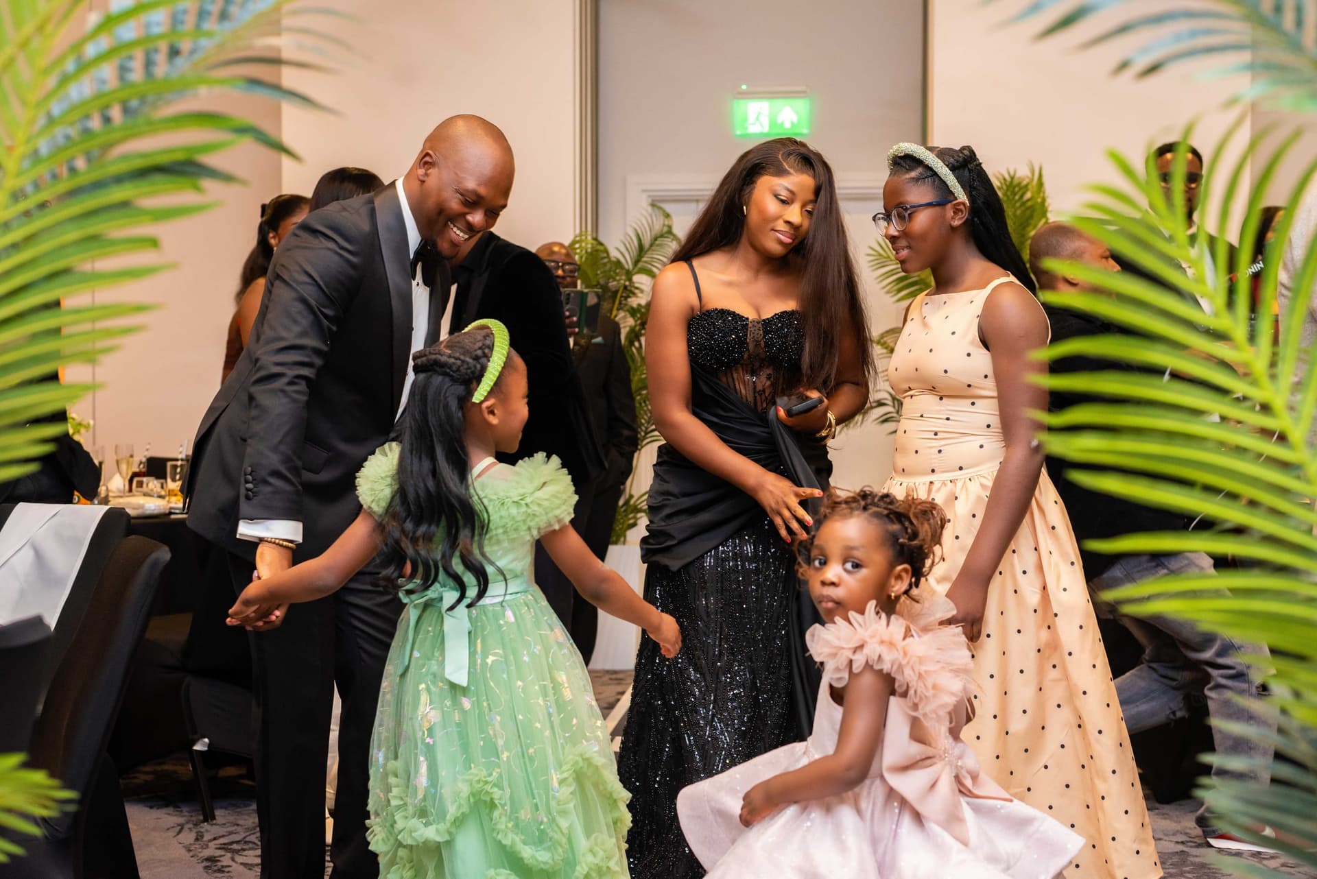 The children come through the room ahead of him — one in green tulle, one in pink — with him leaning down to them and the rest of the family alongside.