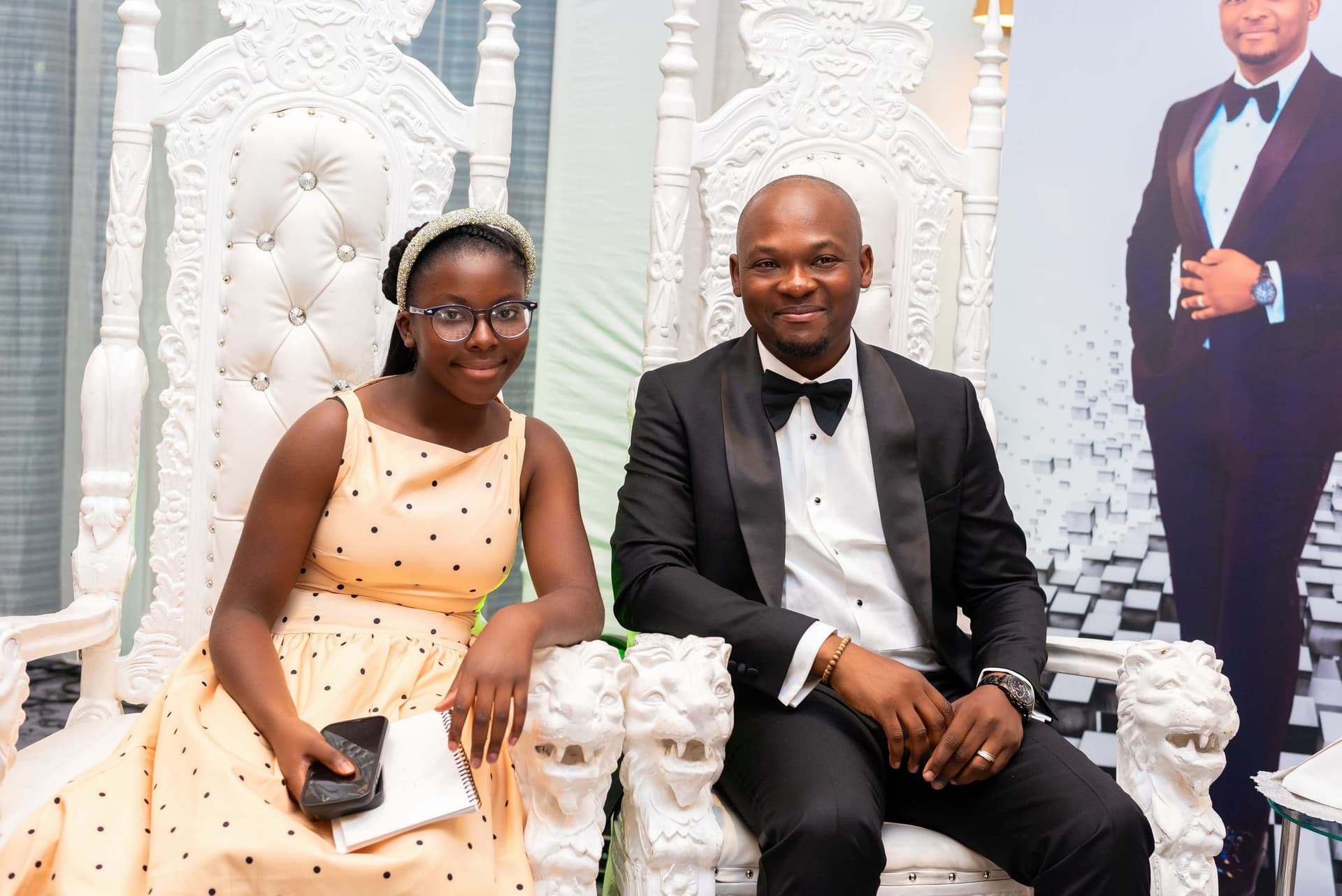 Father and daughter on the thrones, both turned to the camera, the printed board of chess pieces behind them.