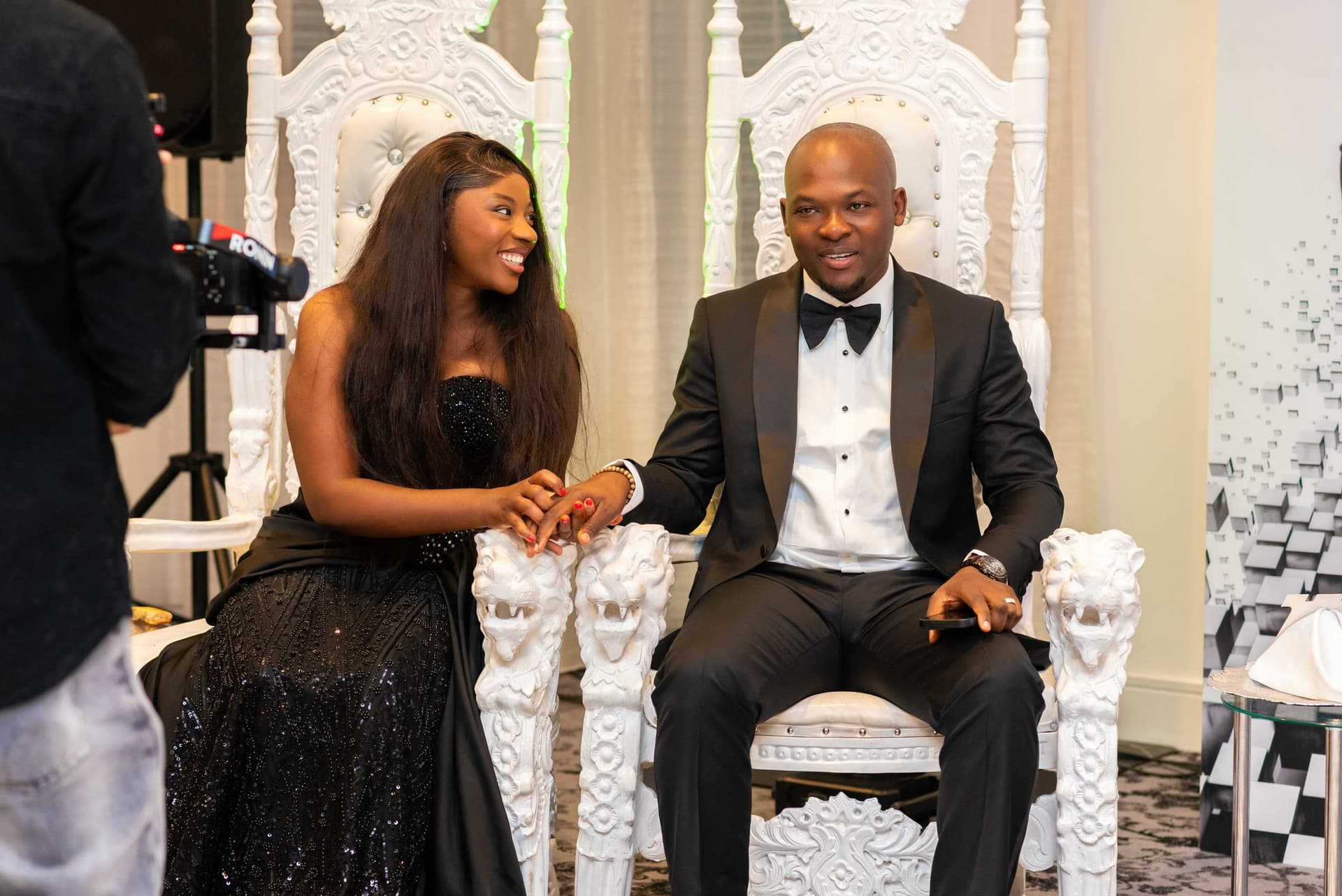 His wife reaches across from the next throne to hold his hand, laughing, a videographer working at the edge of the frame.