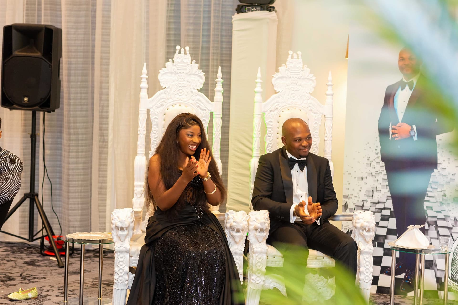 The two of them applauding from the thrones, shot through foreground foliage, a speaker stack and his portrait board behind.