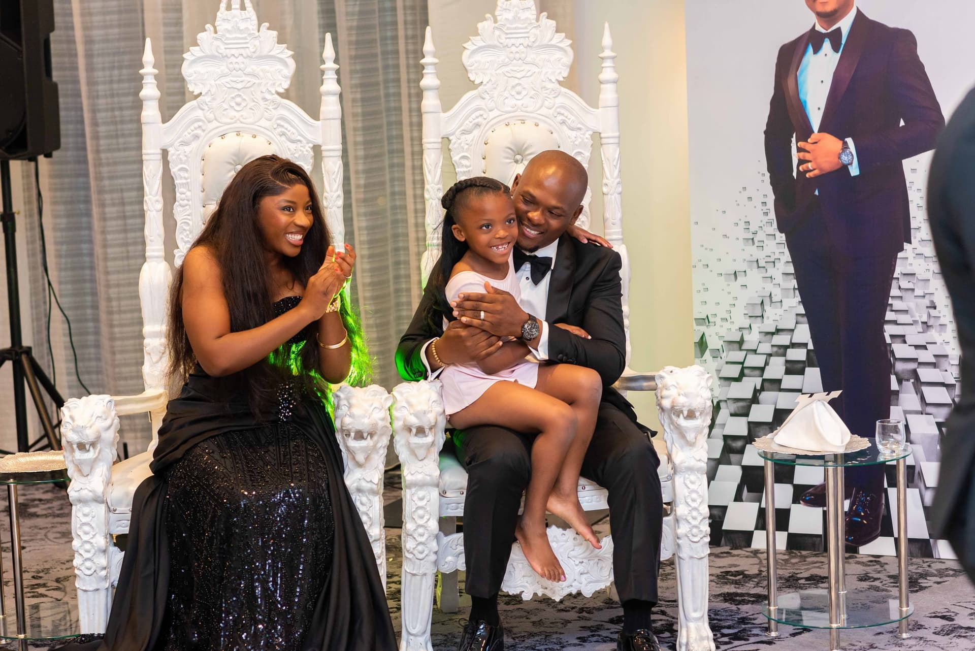 Their youngest climbs into his lap on the throne while his wife claps beside them, all three mid-laugh.