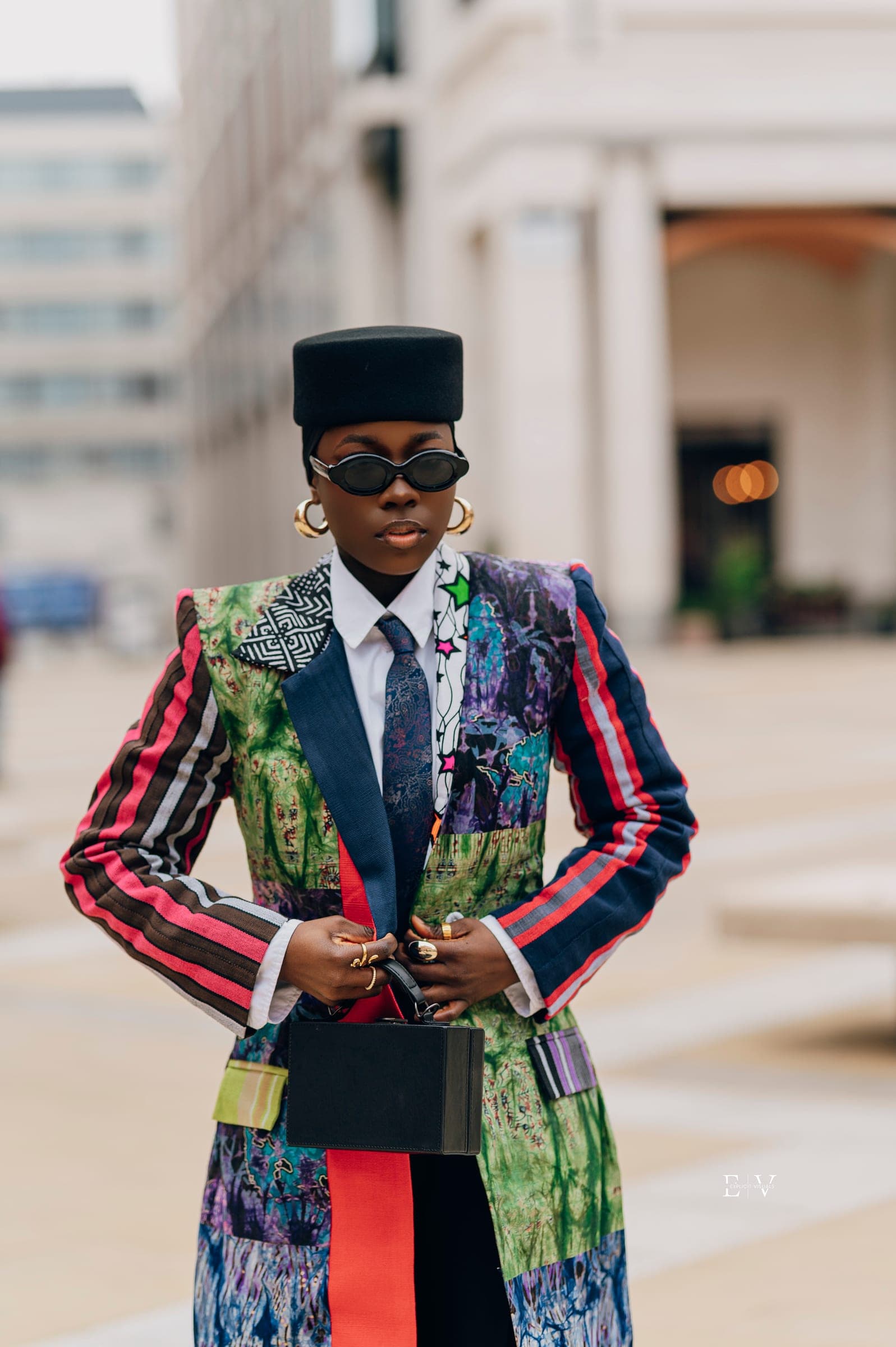 Three-quarter length, hands on hips, the full patchwork coat visible — green, blue and printed panels split by a red vertical stripe, a black box bag held at the waist.