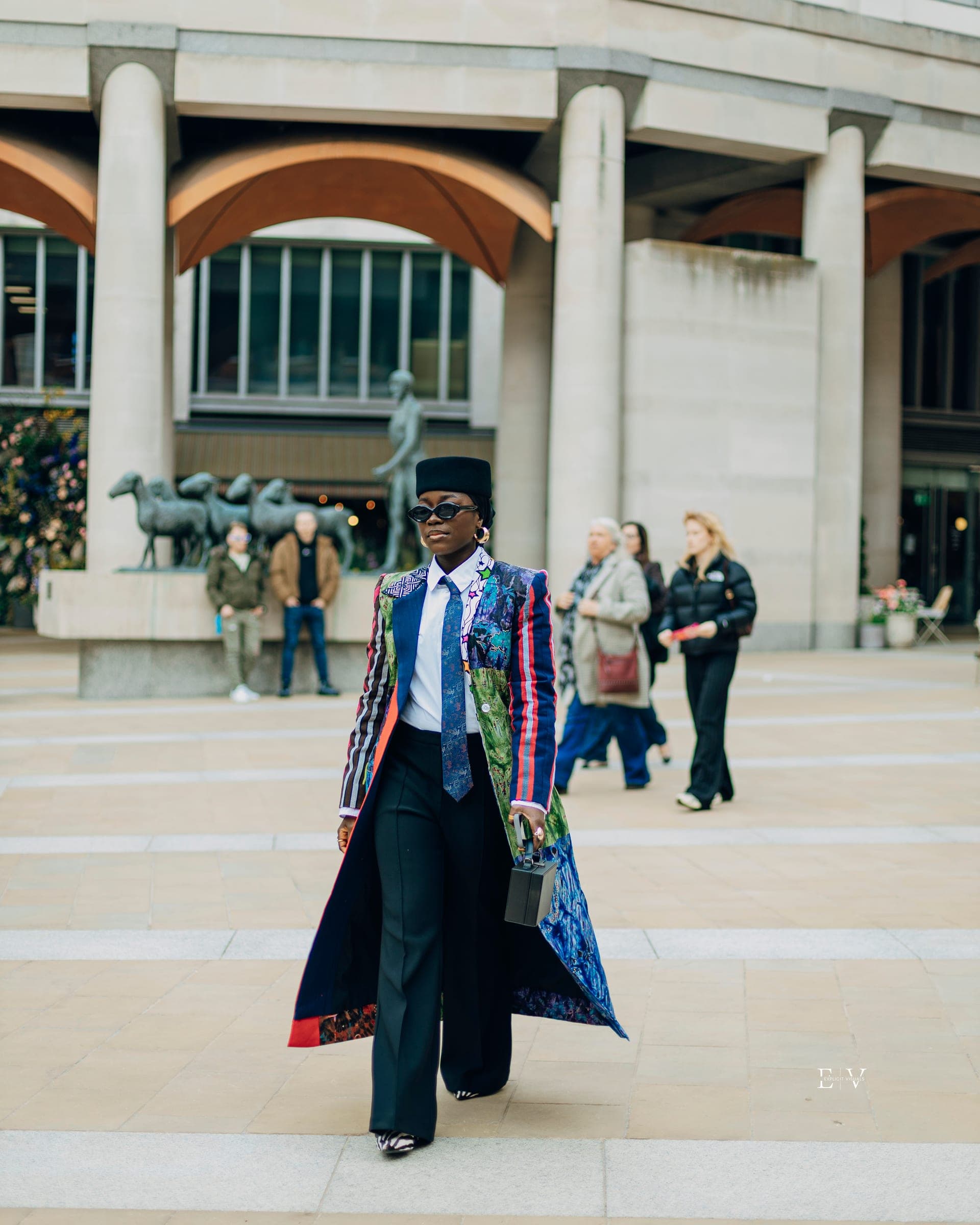 A woman in a black hat, sunglasses and a long printed coat over a white shirt and black trousers, walking toward the camera across a paved square with a colonnade and bronze animal sculptures behind her.