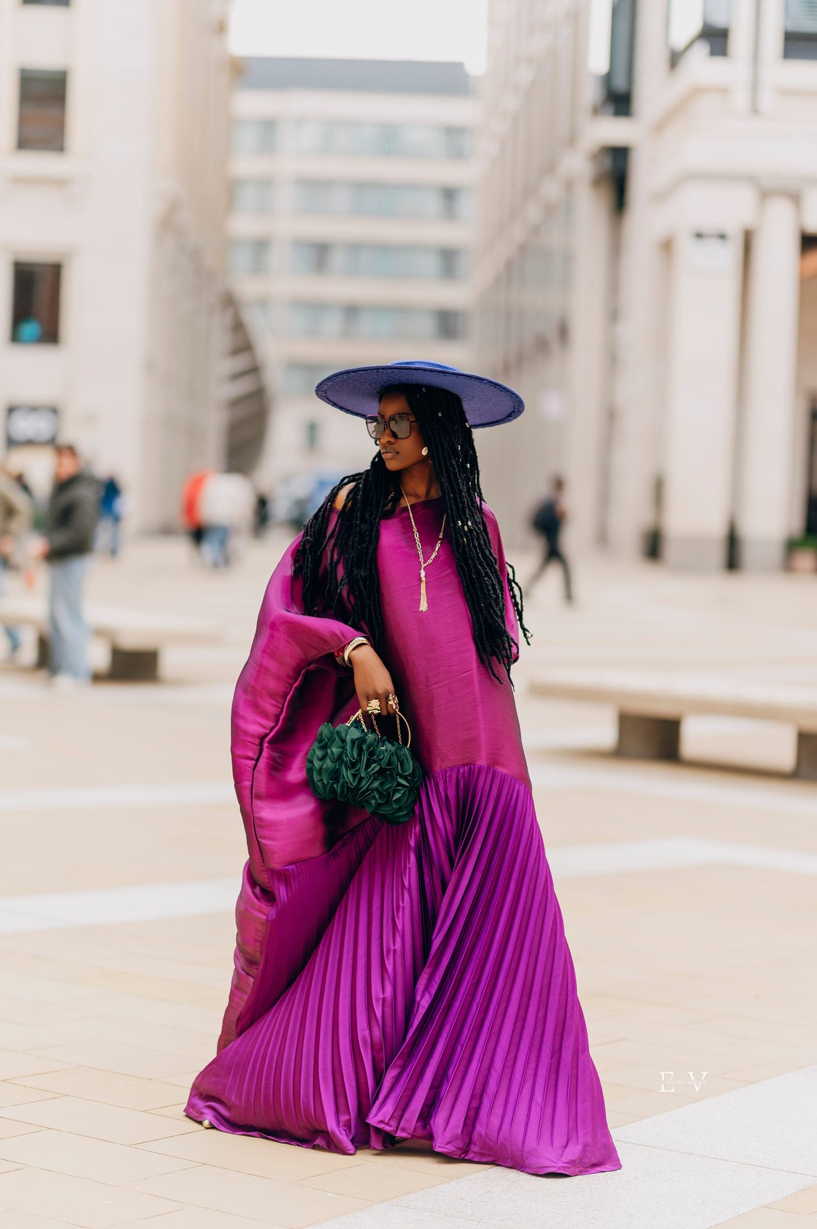 The same gown standing still, the satin cape falling from one shoulder, tall pale office buildings soft behind her.