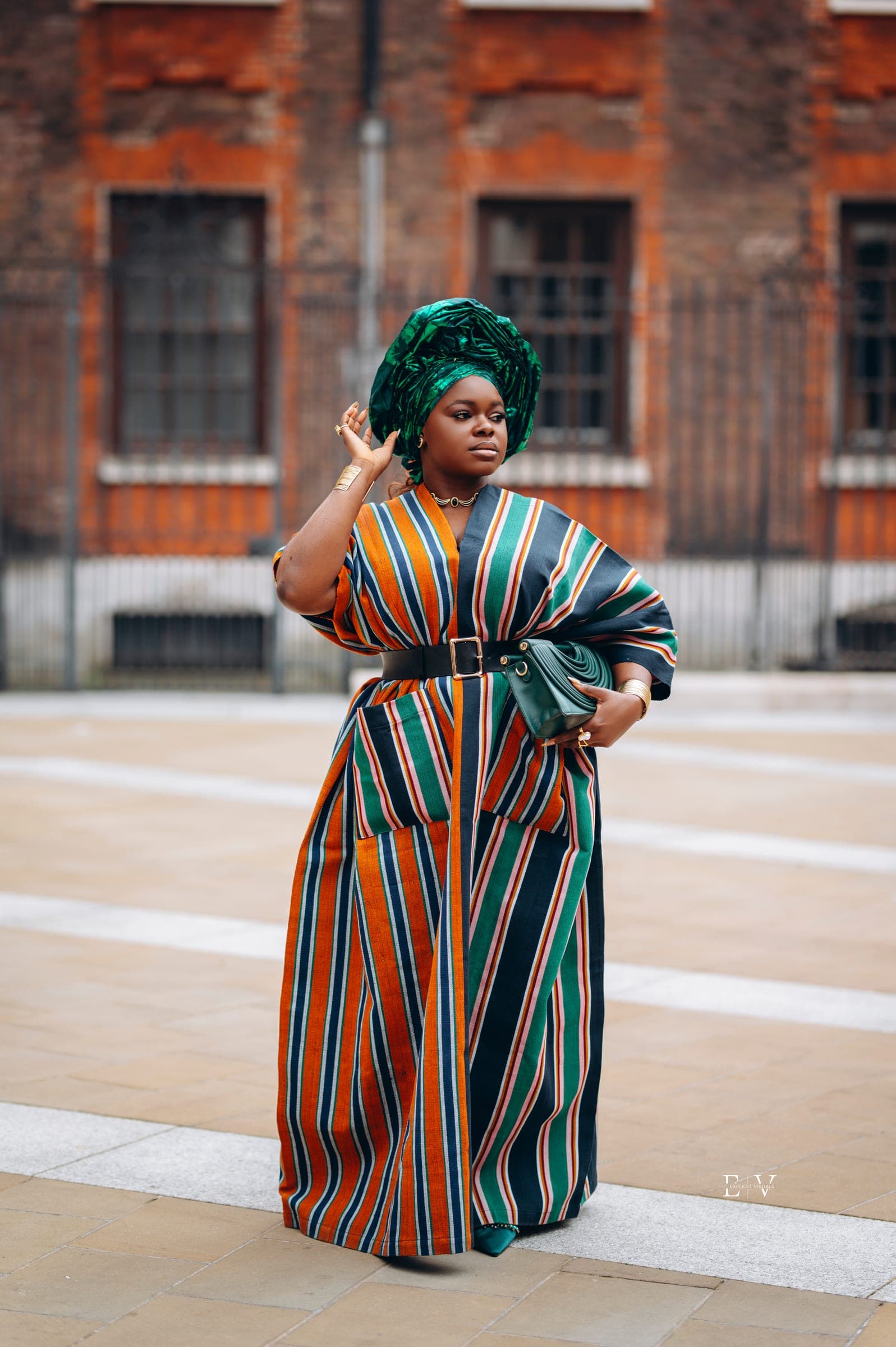 A woman in a green gele and an orange, green and navy striped gown, one hand raised toward her head, a red brick building out of focus behind her.